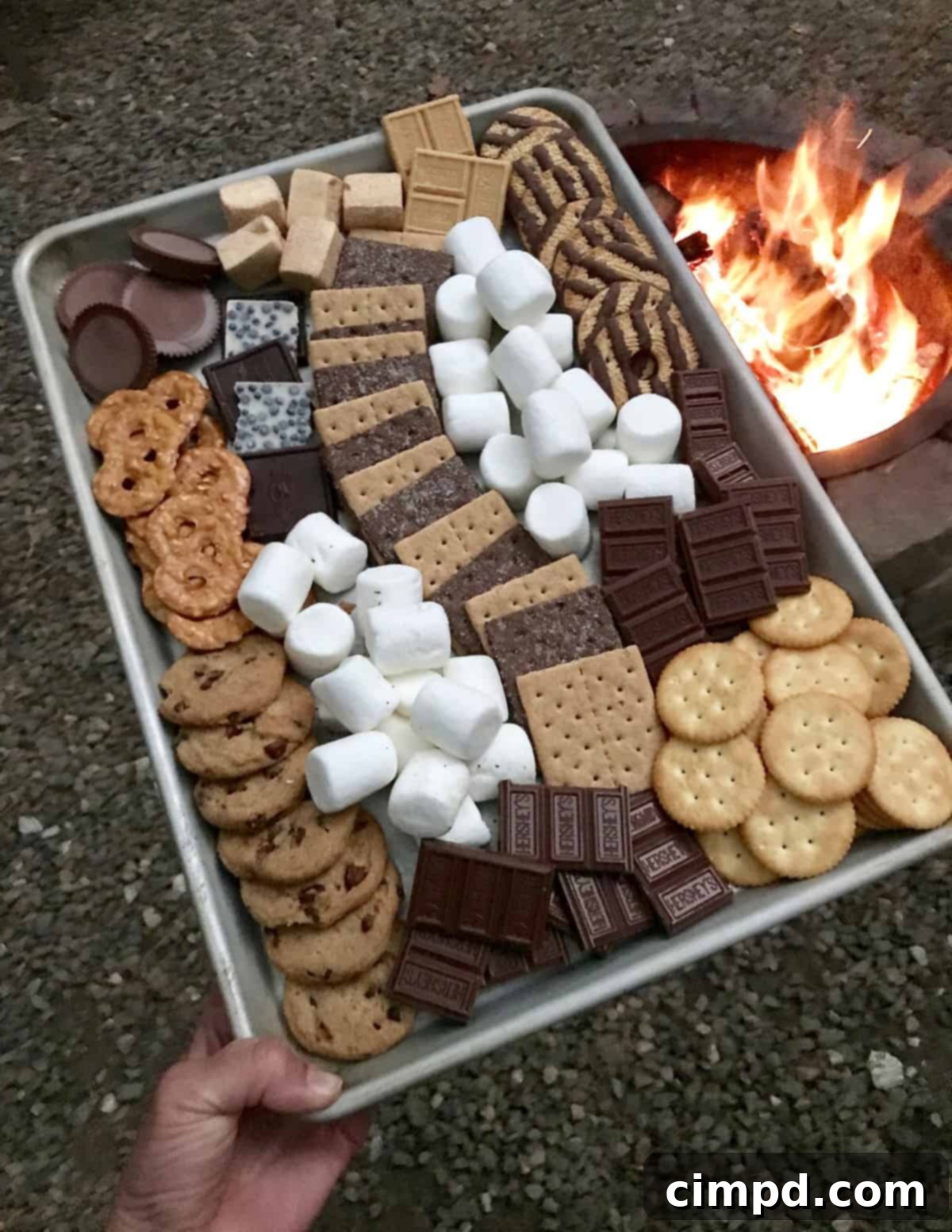 A hand holding a toasted marshmallow over a S'mores Tray, with various ingredients awaiting customization.