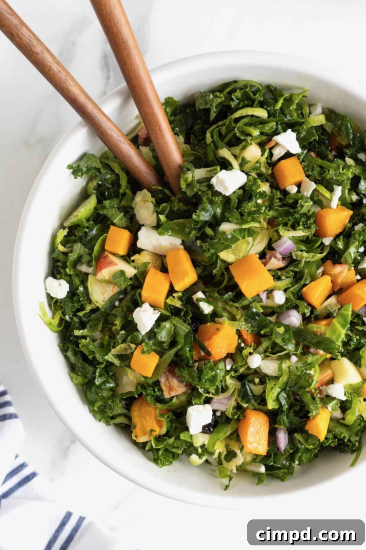 Vibrant Kale and Brussels Sprouts Salad 5 A large white serving bowl of kale salad with wooden salad spoons sticking out.