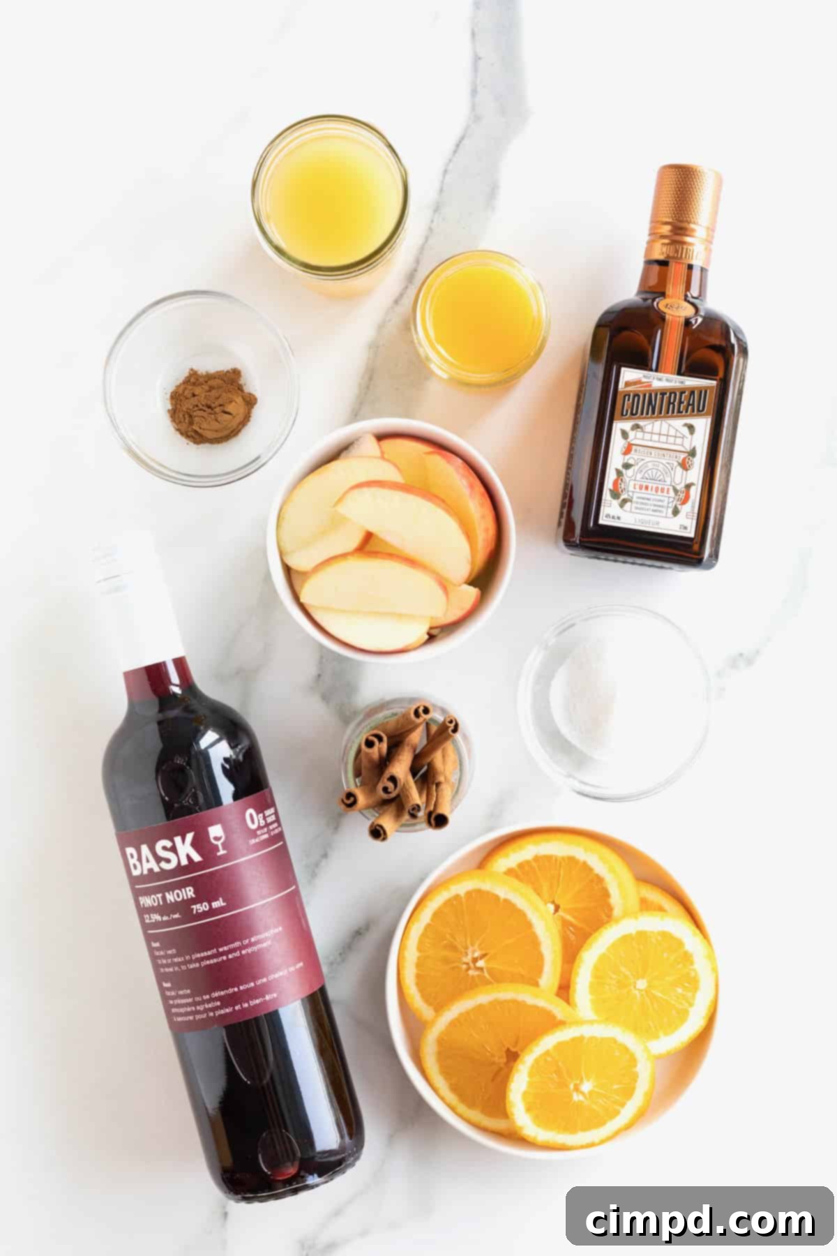 Spiced Apple Cider Sangria 6 Various ingredients for making Apple Cider Sangria, including apples, oranges, apple cider, red wine, and orange liqueur, neatly arranged on a white marble counter.