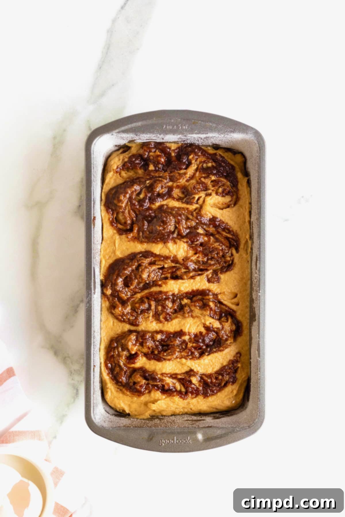 The final cinnamon swirl being artfully applied to the top layer of pumpkin bread batter in an aluminum loaf pan before baking.