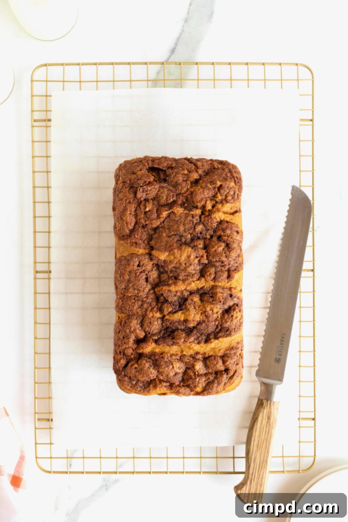 A freshly baked loaf of cinnamon swirled pumpkin bread on a parchment-lined cooling rack, next to a bread knife.