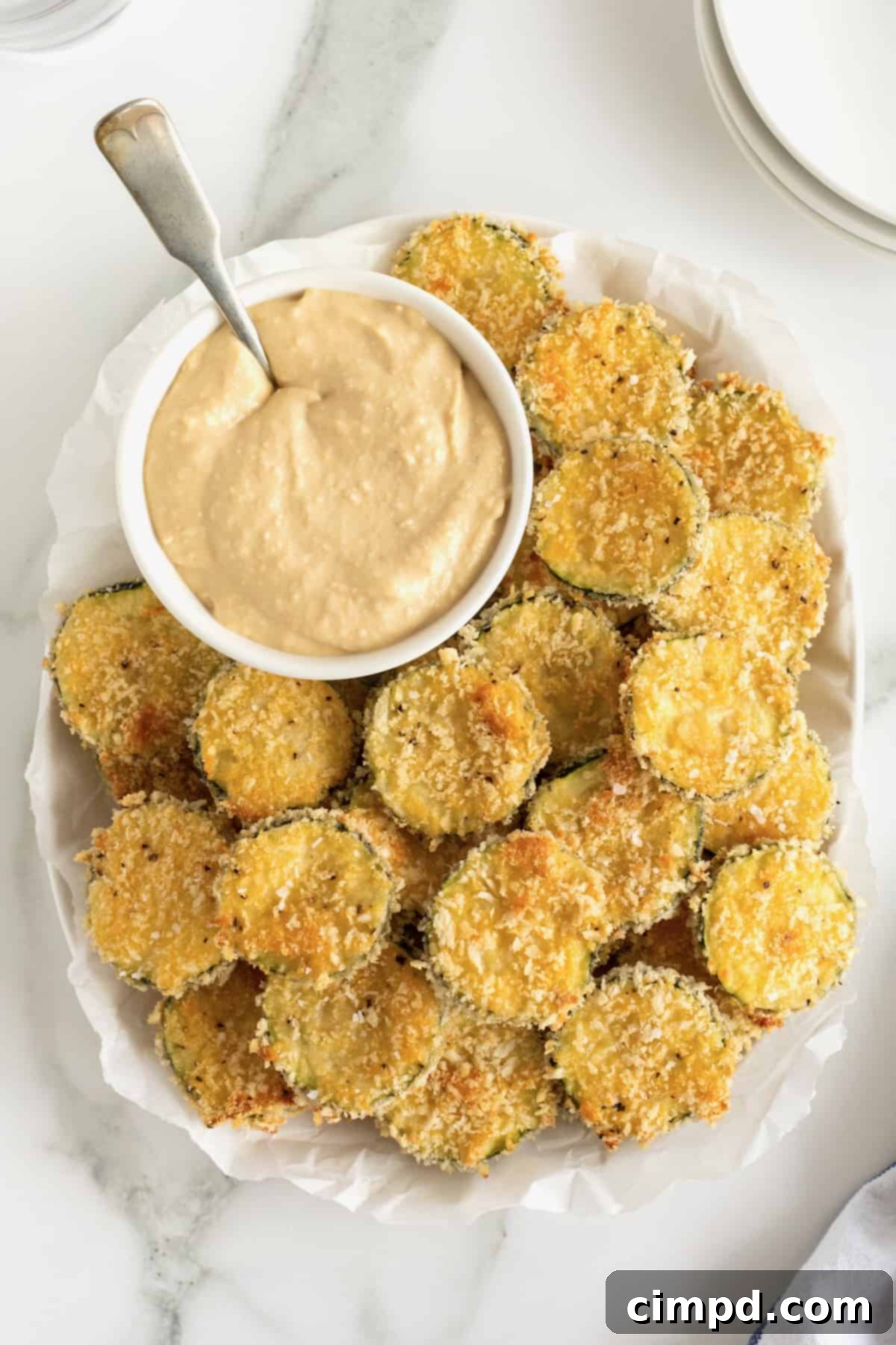 Crispy Zucchini Snacks 2 An oblong white serving platter of zucchini chips surrounding a bowl of dipping sauce with a metal spoon in it.
