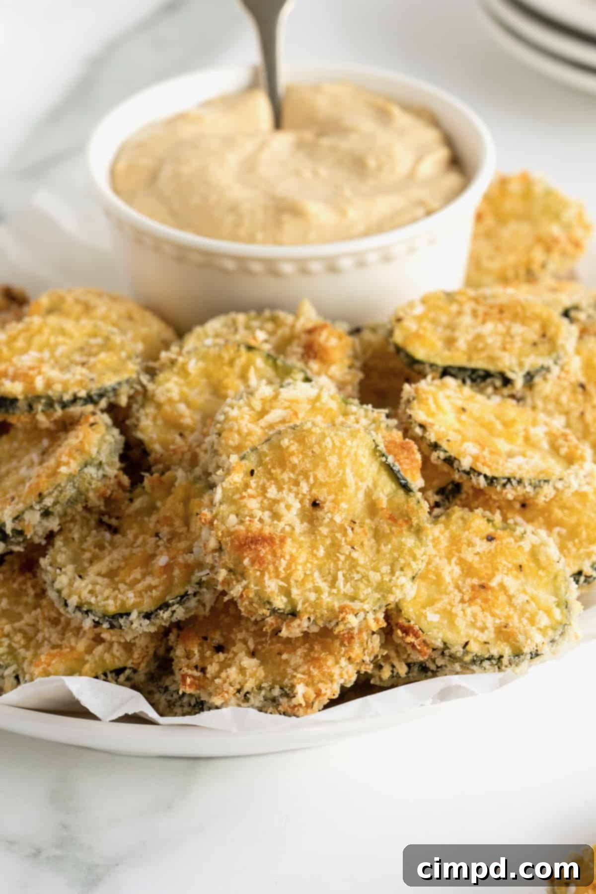 Crispy Zucchini Snacks 5 An oblong white serving platter of zucchini chips surrounding a bowl of hummus.