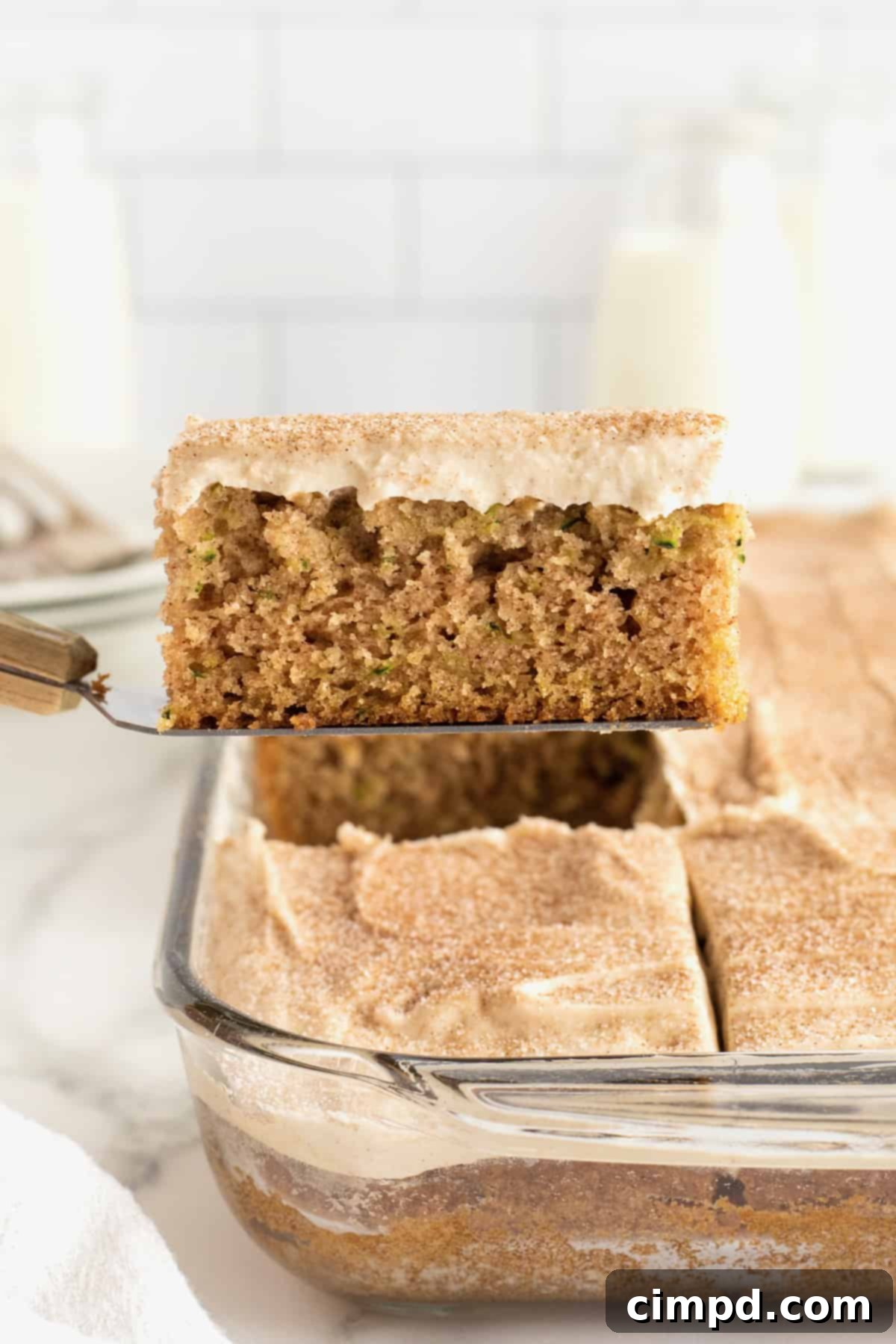 A generous slice of zucchini cake, frosted with cinnamon cream cheese frosting, being lifted from the baking dish with a spatula.