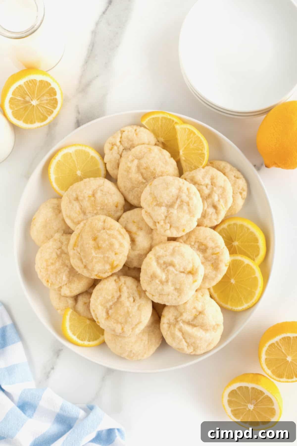 A large white round serving plate generously piled with golden lemon cookies, each dusted with sugar. Several vibrant lemon slices are arranged artistically around the cookies, adding a fresh garnish to the presentation.
