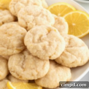 A large white round serving plate piled with lemon cookies dusted with sugar. There are two lemon slices garnishing the plate on the upper right hand.