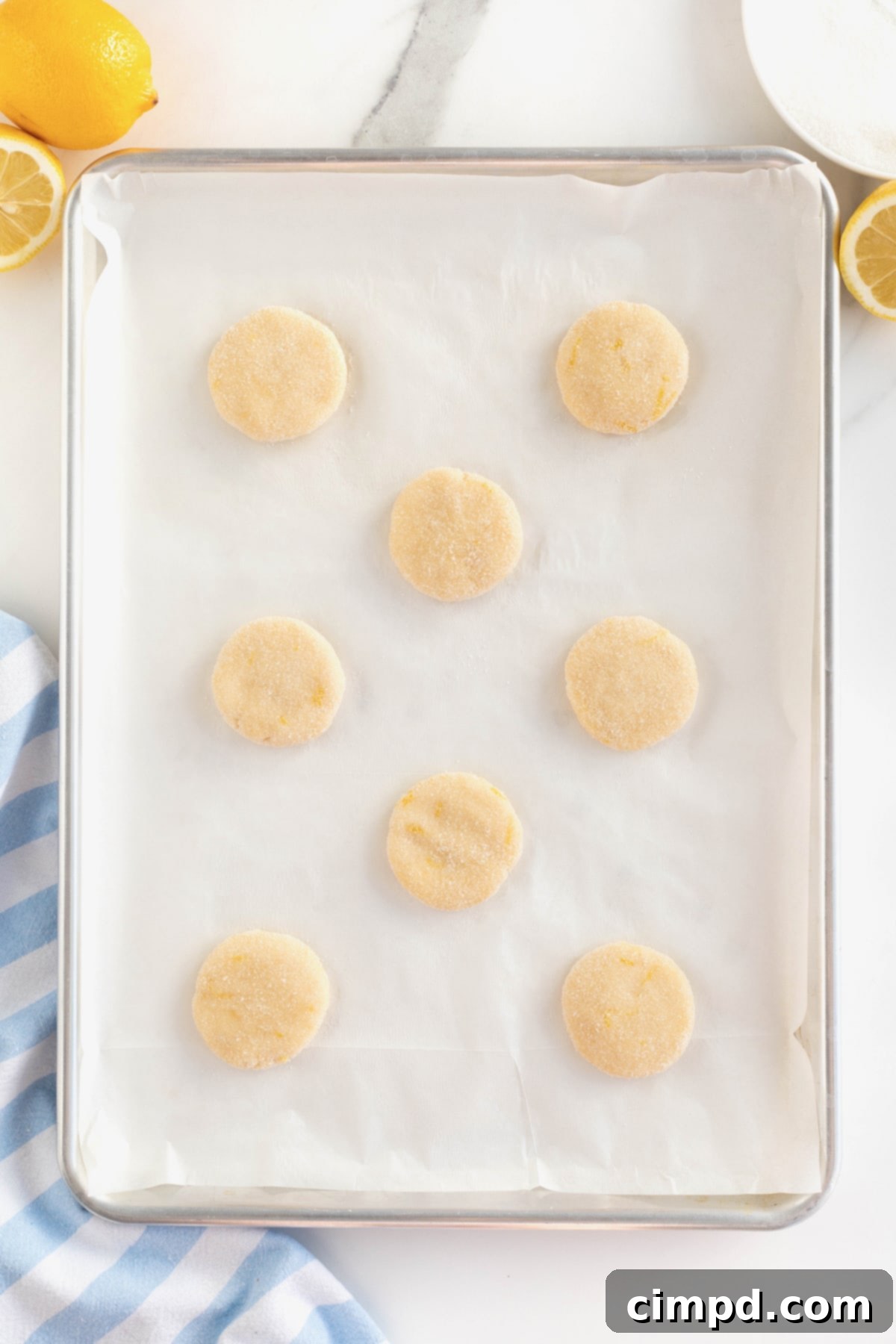 Eight un-baked, flattened lemon cookies are neatly arranged on a parchment-lined aluminum baking sheet, perfectly spaced for baking.