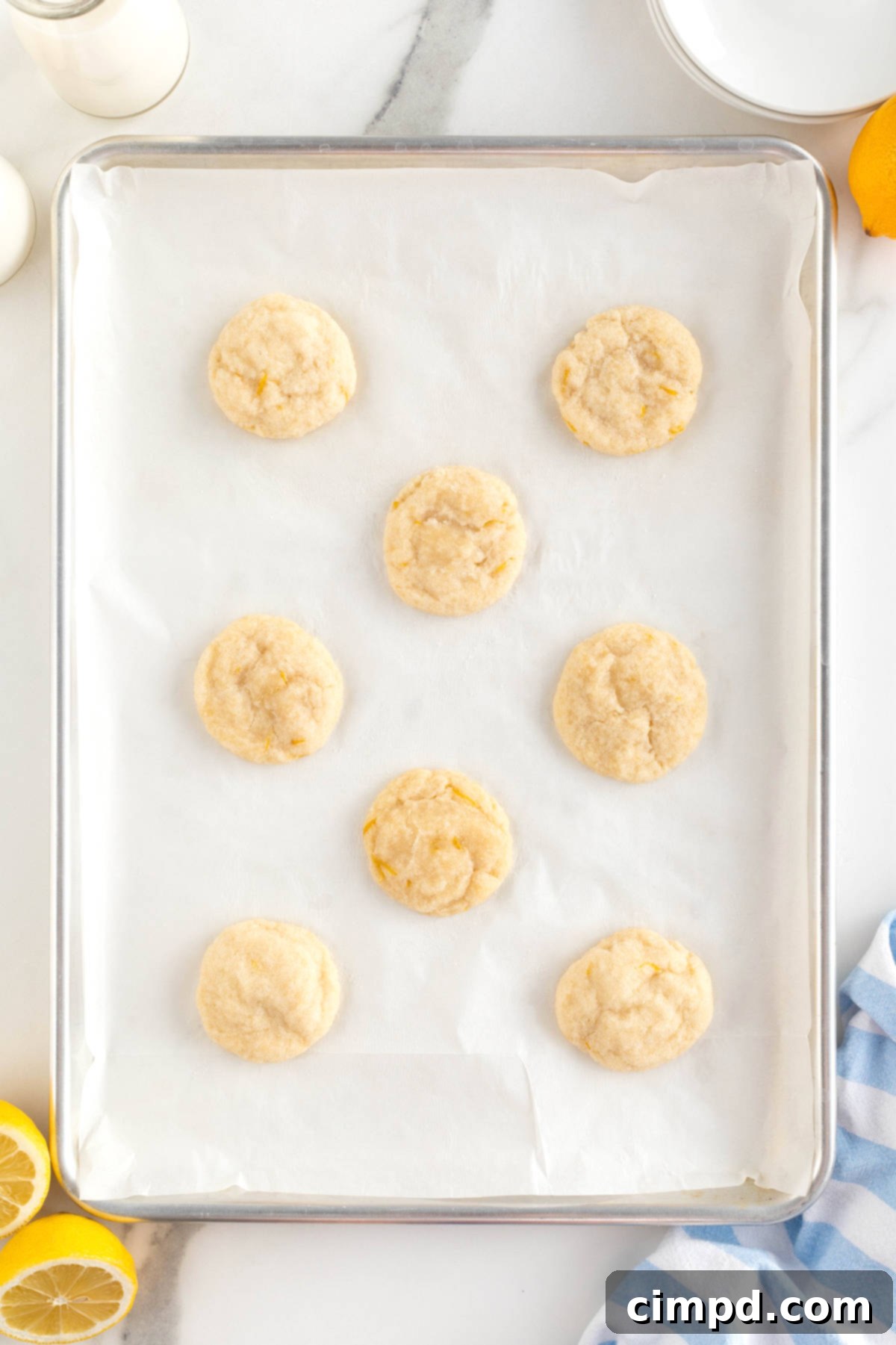 Eight freshly baked lemon cookies, with slightly golden edges, are shown cooling on a parchment-lined aluminum baking sheet, still warm from the oven.
