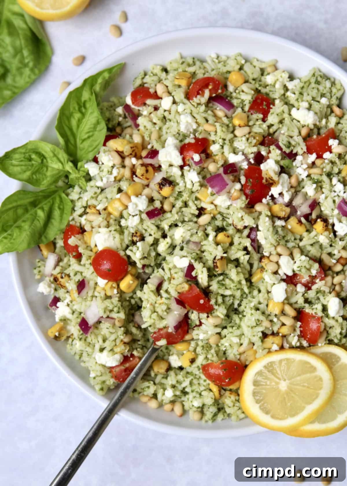A vibrant Pesto Rice Salad in a white bowl, garnished with fresh basil leaves, roasted corn, and cherry tomatoes, against a rustic wooden background.