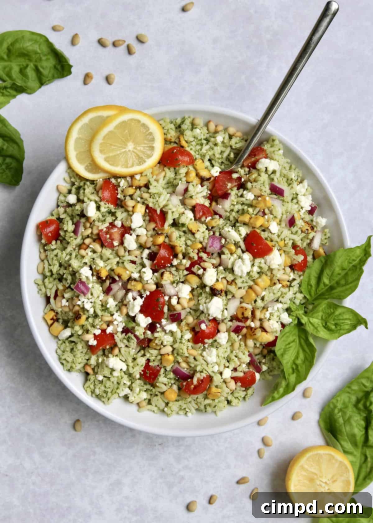 A close-up shot of a serving of Pesto Rice Salad in a white bowl, garnished with fresh basil, roasted corn, and goat cheese crumbles, ready to be enjoyed.