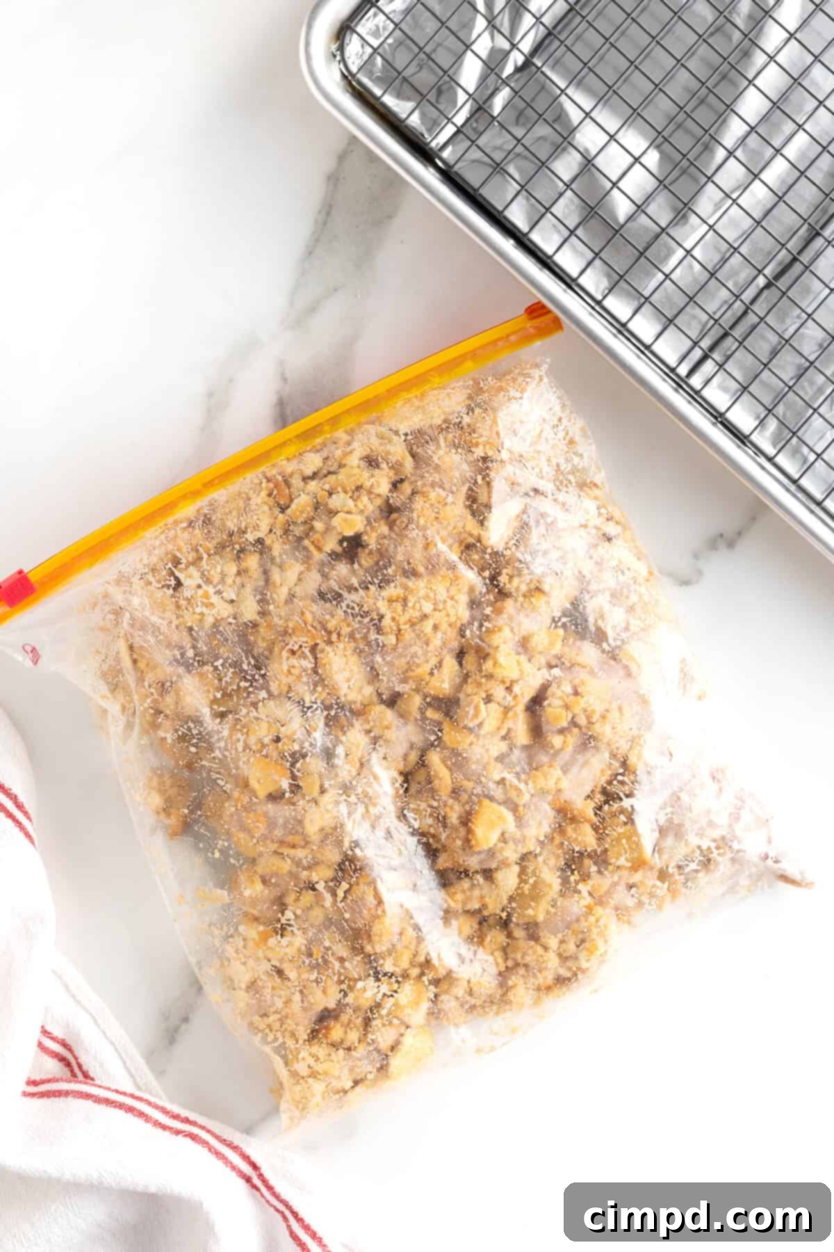 A resealable plastic bag containing chicken pieces thoroughly coated in Ritz cracker crumbs, resting on a white marble counter.