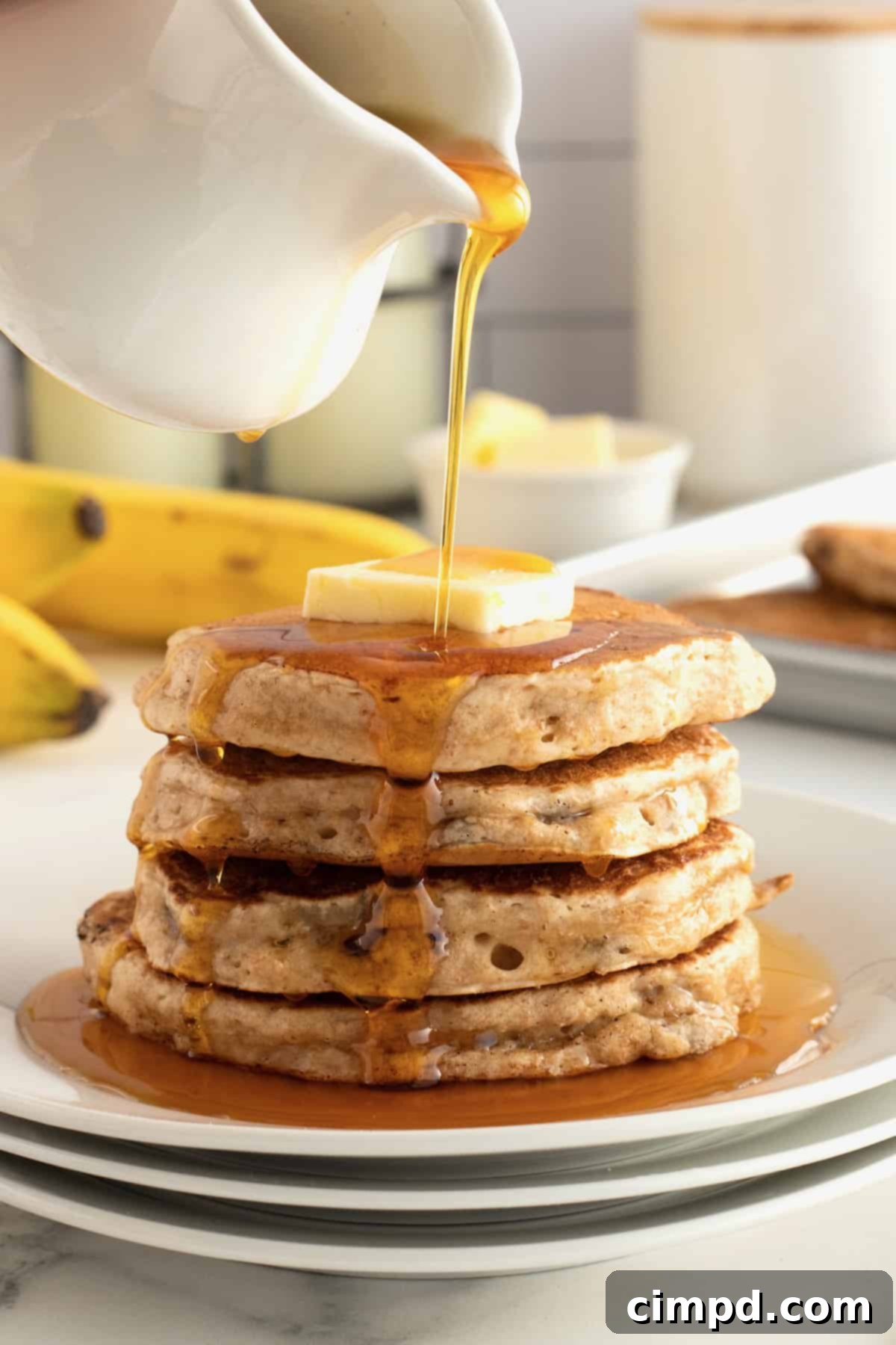 A stack of golden brown Whole Wheat Banana Pancakes by The BakerMama, drizzled with maple syrup.
