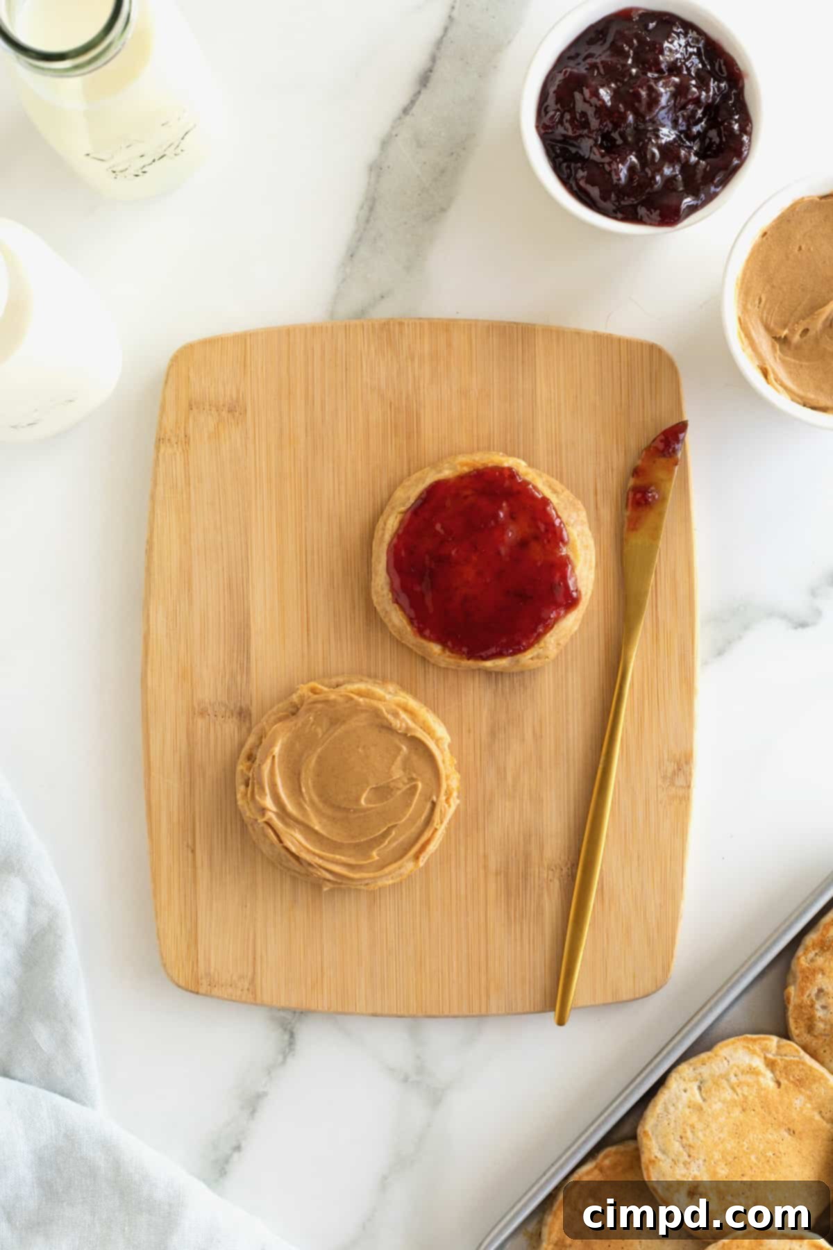 Two banana pancakes on a wood cutting board. One is spread with peanut butter and the other is spread with jelly.