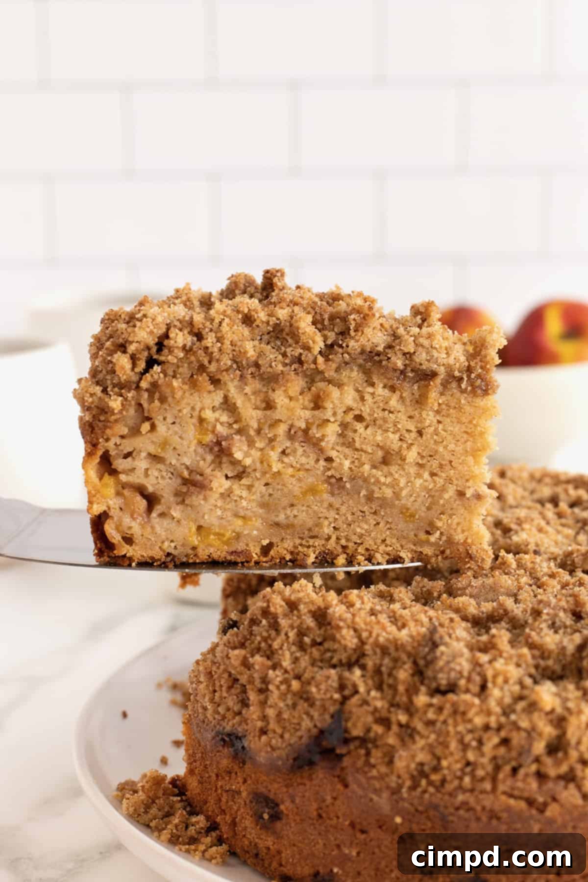 A perfectly sliced piece of peach coffee cake, revealing its moist crumb and pockets of sweet peaches, topped with a generous cinnamon streusel, presented on a pie server with a blurred background of fresh peaches.
