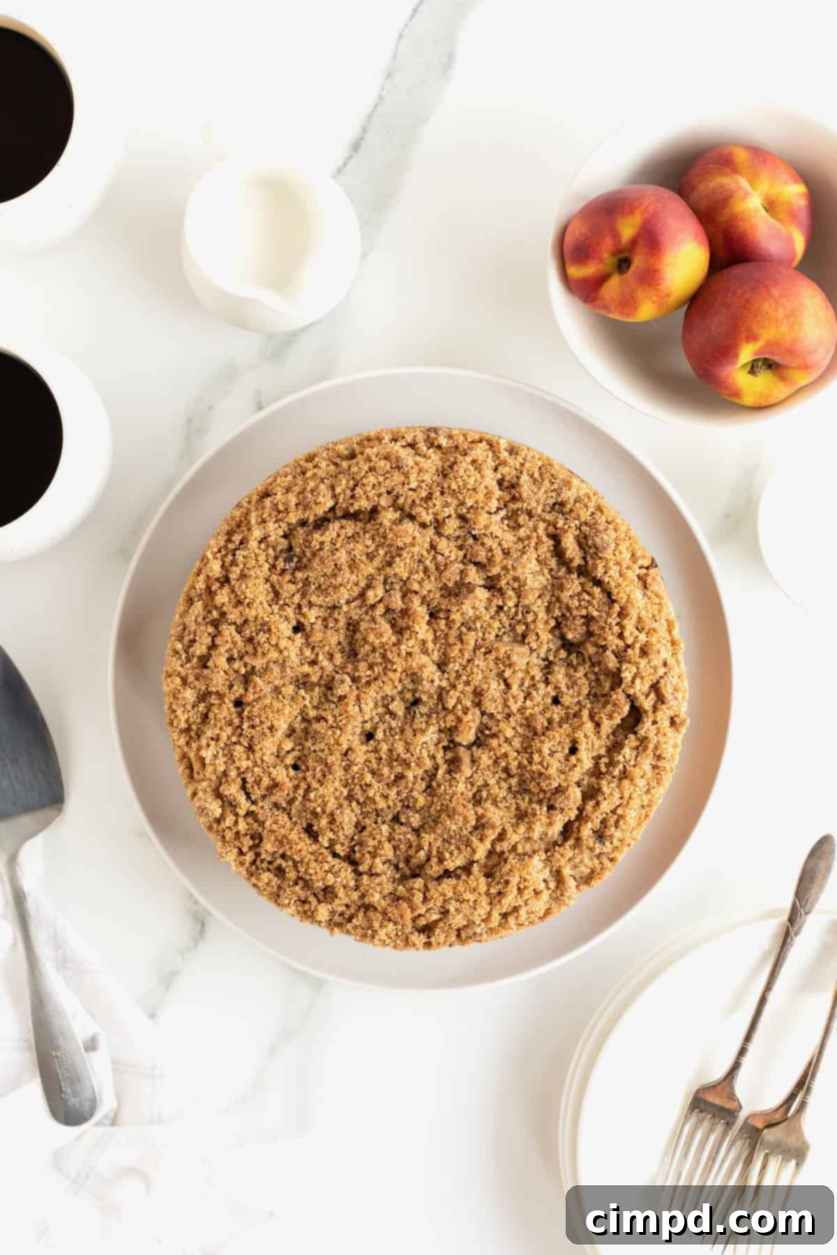 A beautifully baked peach coffee cake with a cinnamon streusel topping, presented on a white serving platter next to a bowl overflowing with fresh, ripe peaches, evoking a sense of abundance.