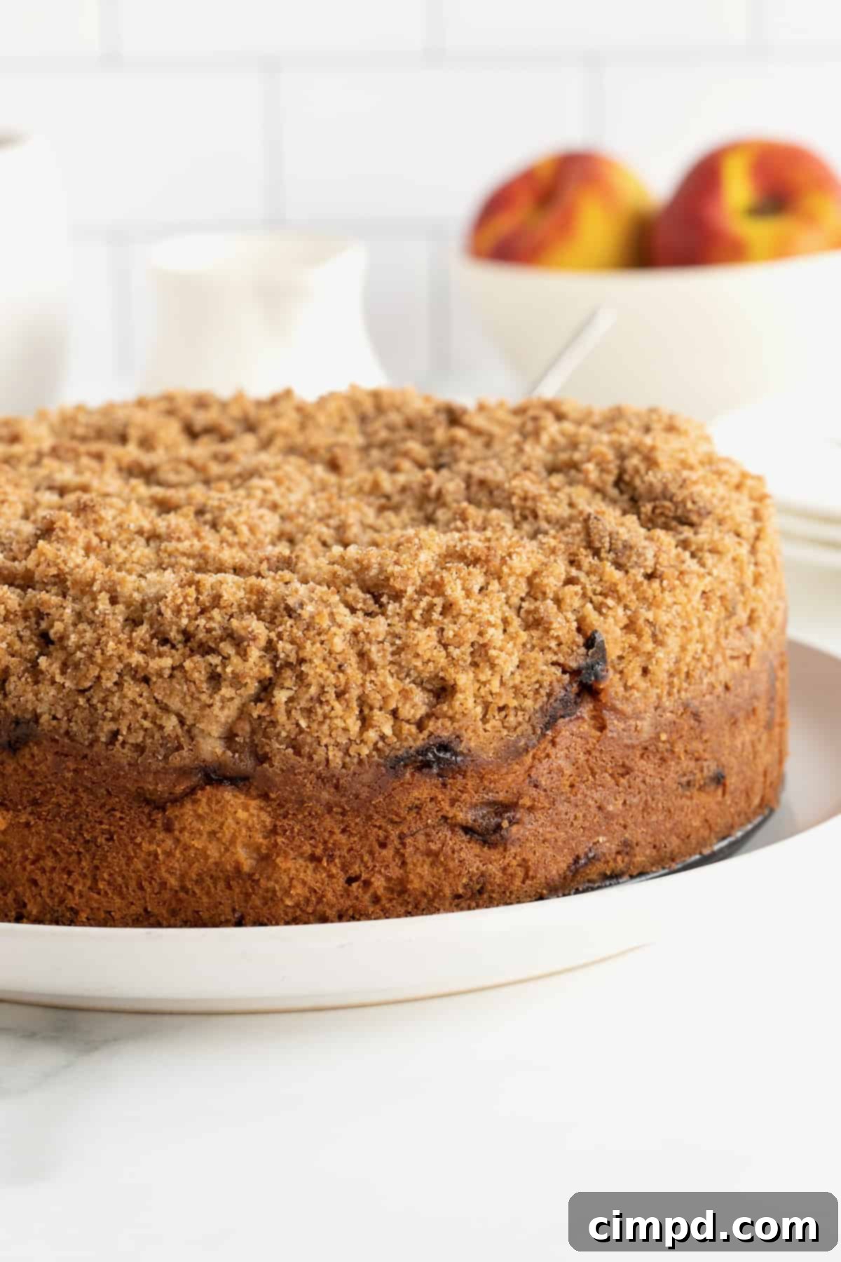 A stunning whole peach coffee cake, beautifully golden brown with a rich cinnamon sugar streusel topping, sitting on a pristine white serving platter, ready to be enjoyed.