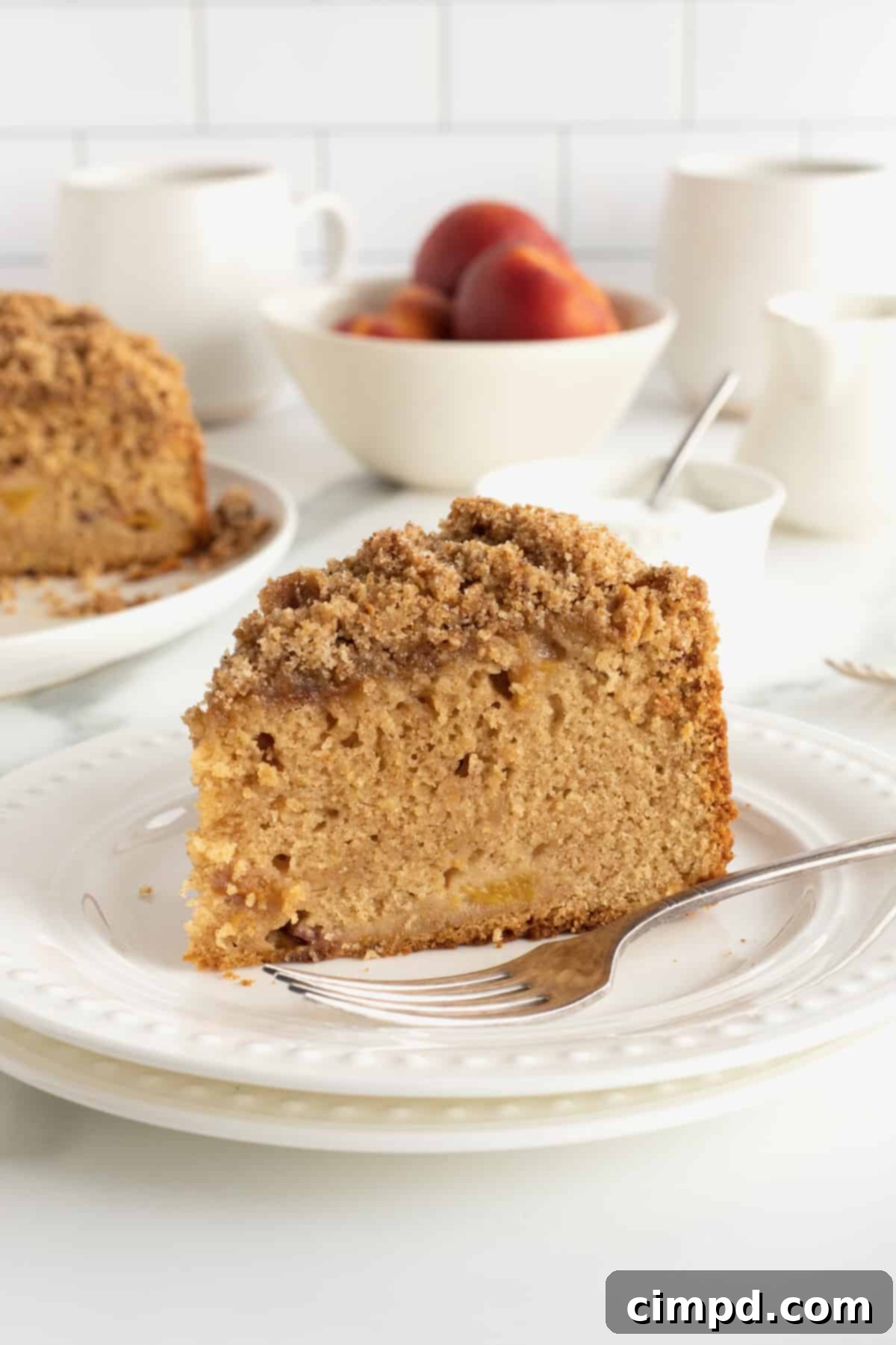 A close-up of a delectable slice of peach coffee cake on a simple white plate, accompanied by a fork, highlighting the generous streusel topping and the tender, peach-filled interior.