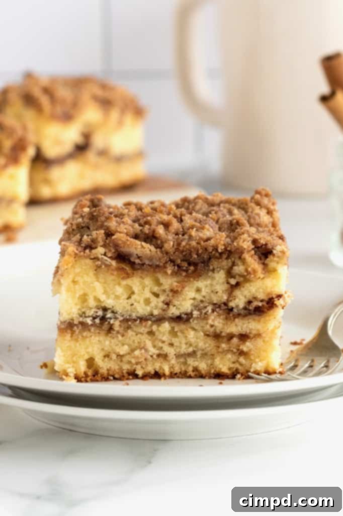 A square slice of classic coffee cake, featuring a golden streusel topping, placed on a stack of white ceramic plates beside a metal fork, emphasizing its inviting texture.