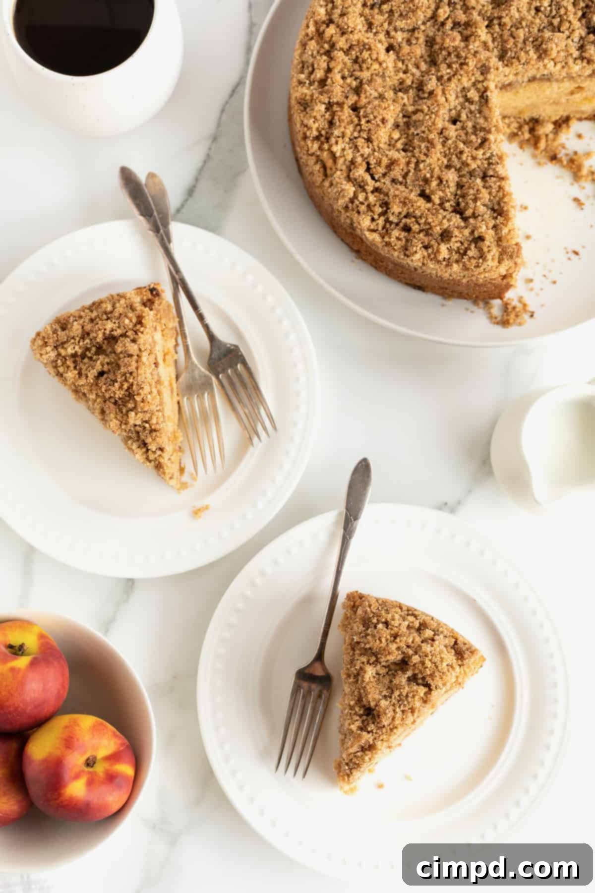 Two enticing slices of peach coffee cake arranged on white plates with forks, with fresh whole peaches positioned in the foreground, creating a vibrant and appealing scene.