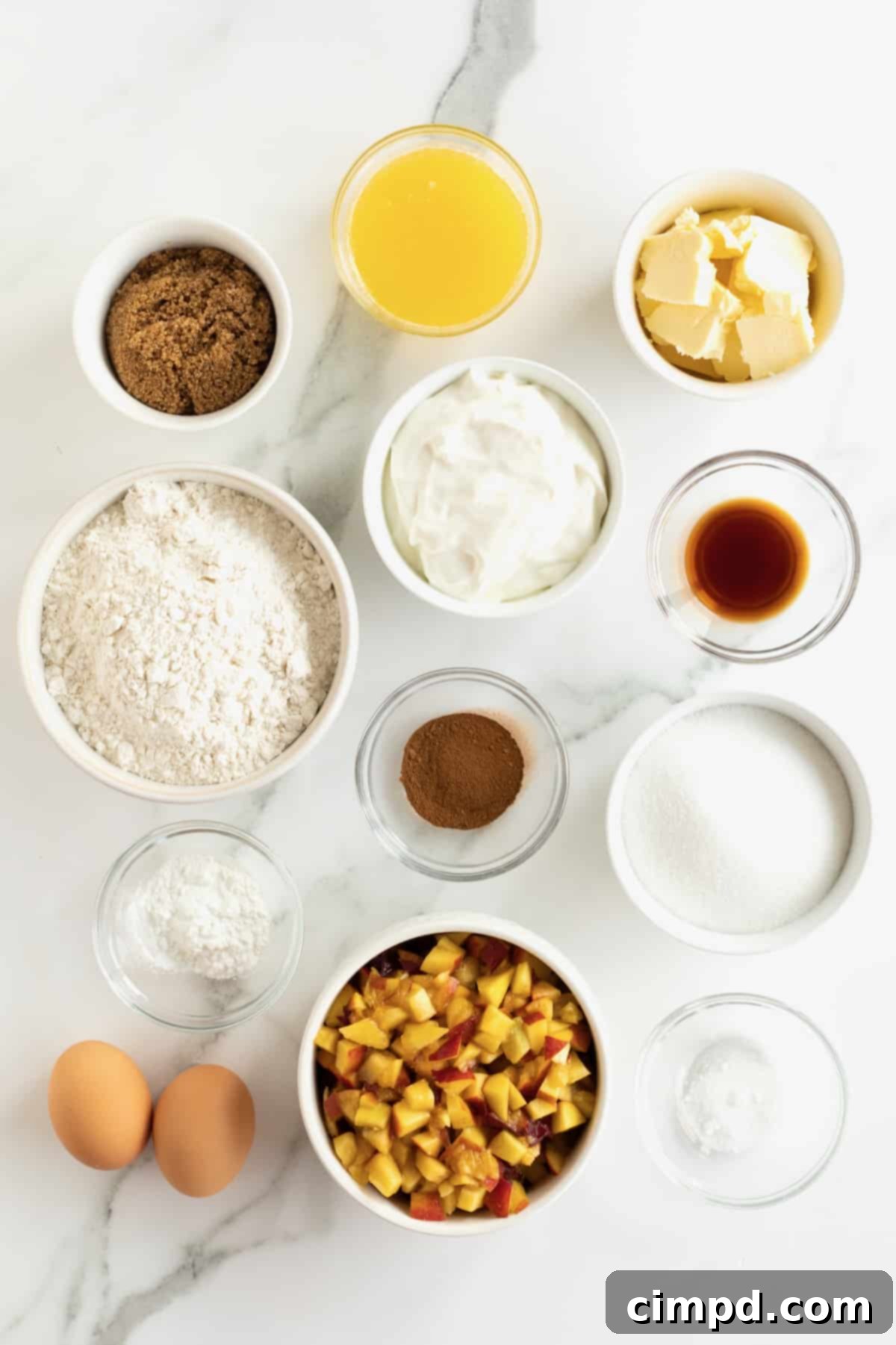 A flat lay photograph showcasing all the individual ingredients for peach coffee cake in small glass dishes on a white marble counter, including butter, sugars, eggs, sour cream, vanilla, flour, baking powder, salt, cinnamon, and chopped fresh peaches.