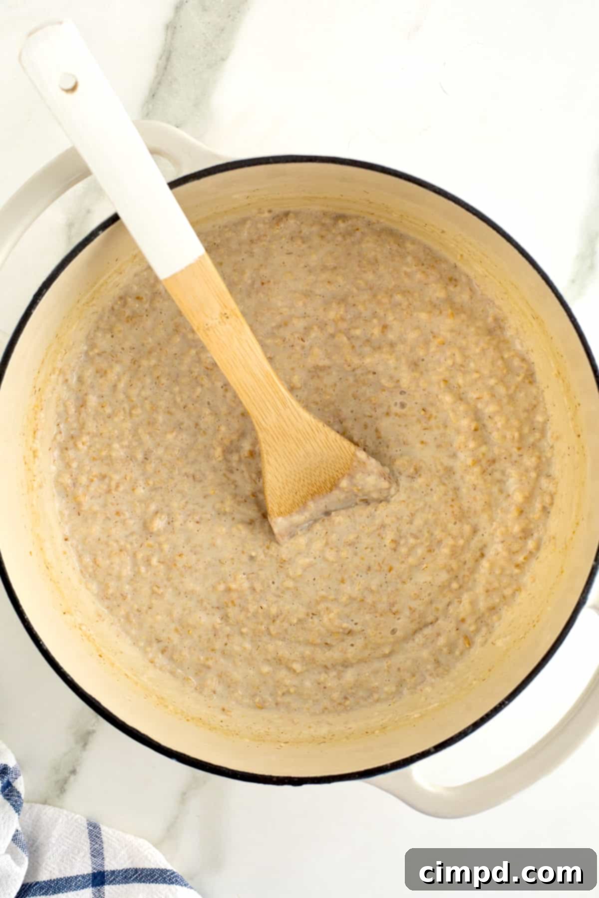 Perfect Steel Cut Oats Every Time 11 A wooden spoon is nestled within a pristine white Dutch oven, full of perfectly cooked, creamy oatmeal, ready to be served and enjoyed.