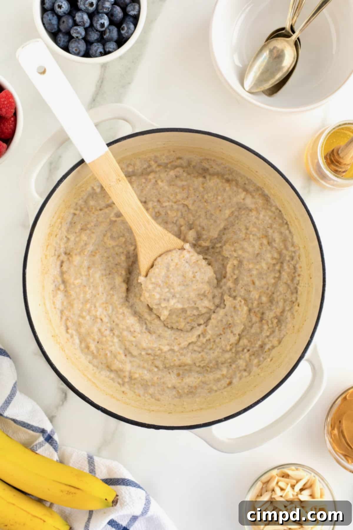 Perfect Steel Cut Oats Every Time 12 A wooden spoon is poised within a pristine white Dutch oven, which holds perfectly reheated, creamy oatmeal, ready for a warm and comforting serving.