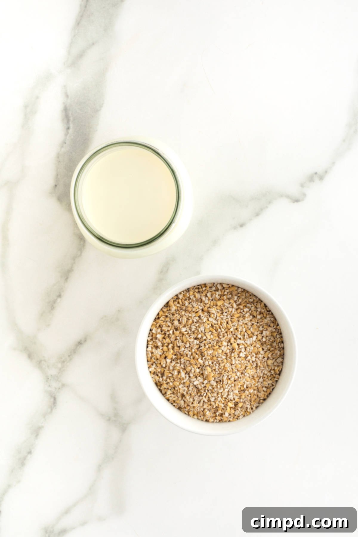 Perfect Steel Cut Oats Every Time 5 A glass jar filled with fresh milk stands beside a pristine white dish brimming with raw steel cut oats, all resting elegantly on a smooth white marble counter, showcasing the simple ingredients for a delicious breakfast.