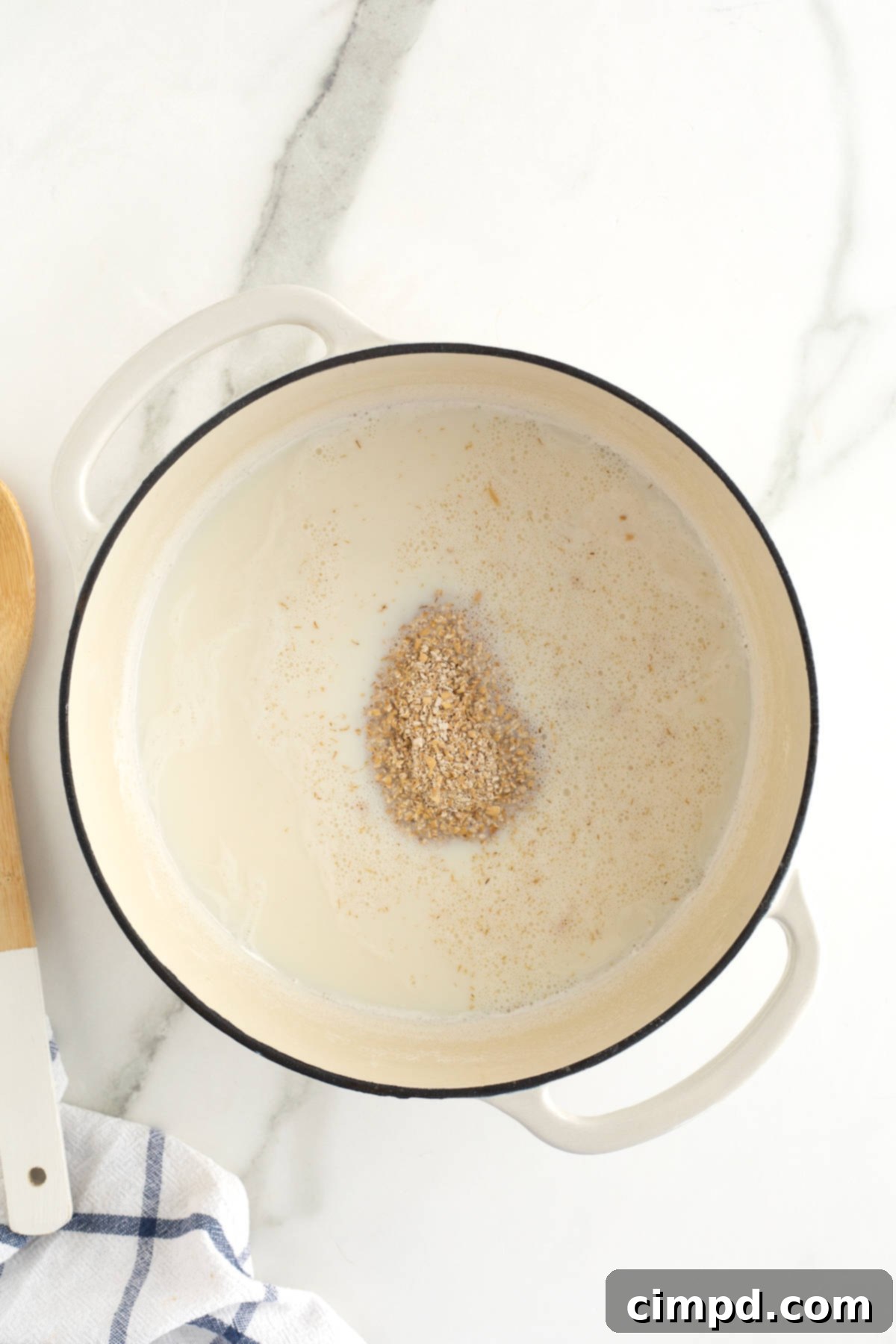 Perfect Steel Cut Oats Every Time 9 A pristine white Dutch oven sits on a stovetop, brimming with hot milk and raw steel cut oats, captured mid-stir as the cooking process begins.