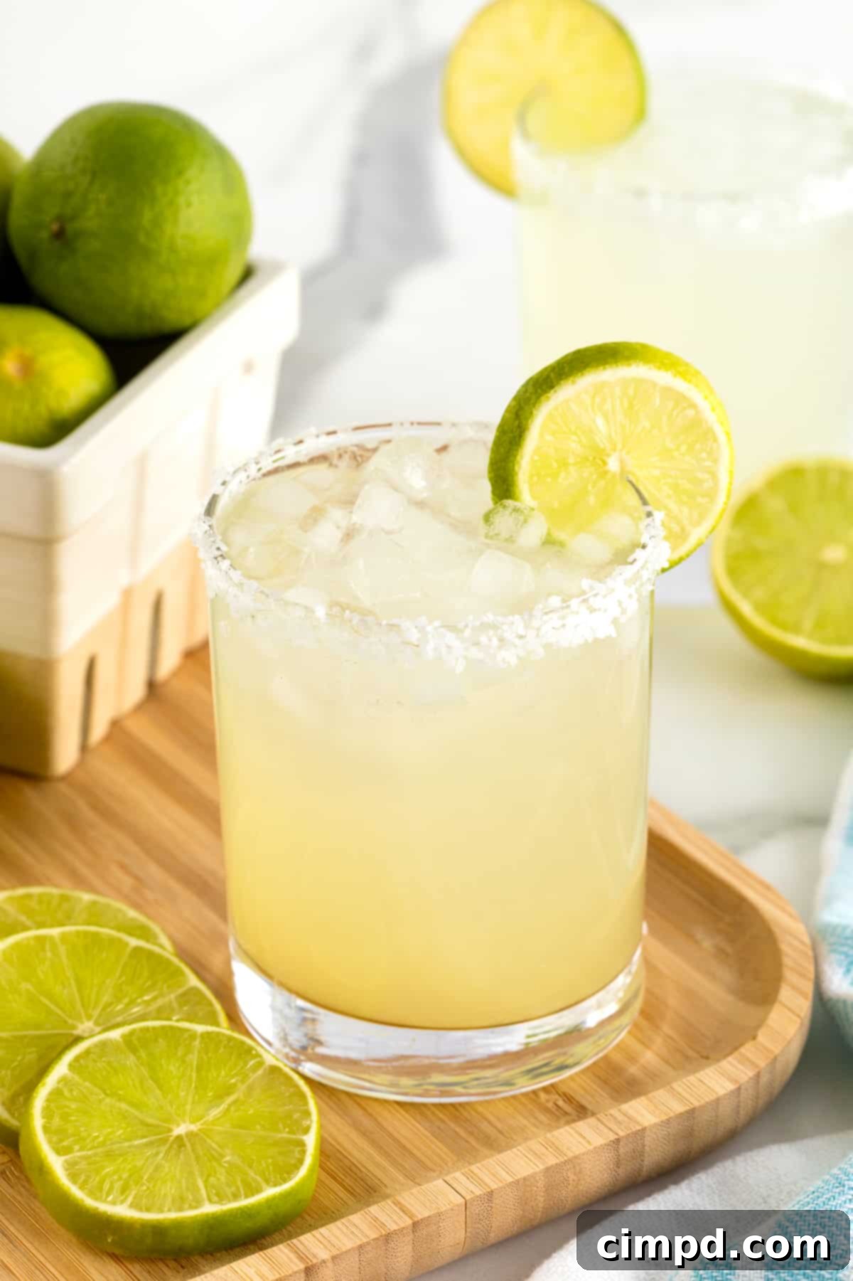A salt rimmed glass filled with margarita on a rimmed wooden serving tray. There is a lime wedge on the rim of the glass.