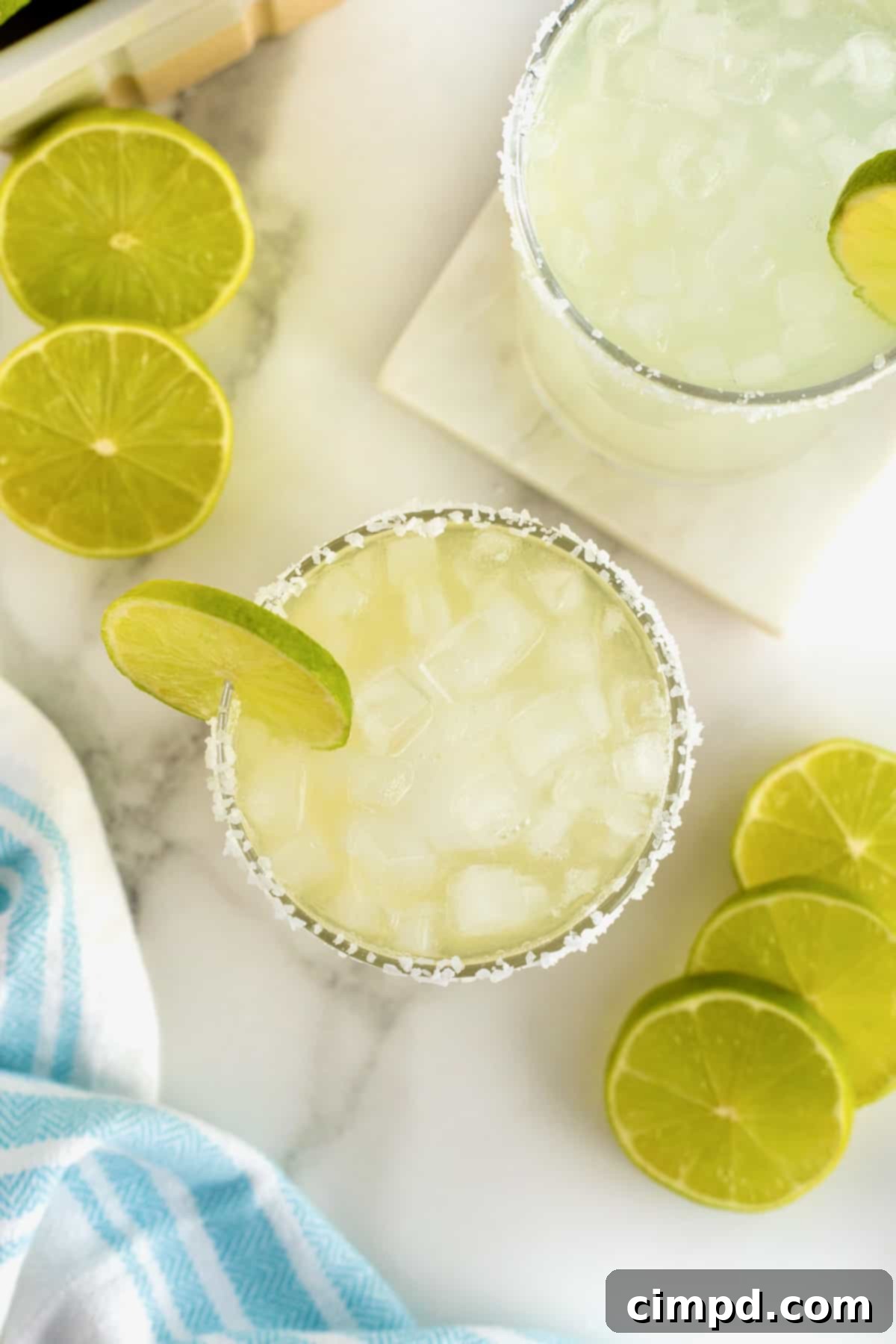 A salt rimmed glass filled with margarita. There is a lime wedge on the rim of the glass and lime slices scattered around.