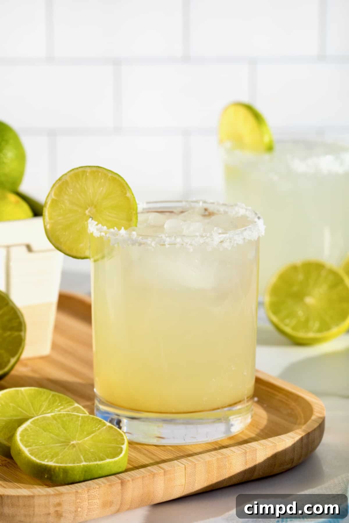 A salt rimmed glass filled with margarita on a rimmed wooden serving tray. There is a lime wedge on the rim of the glass.
