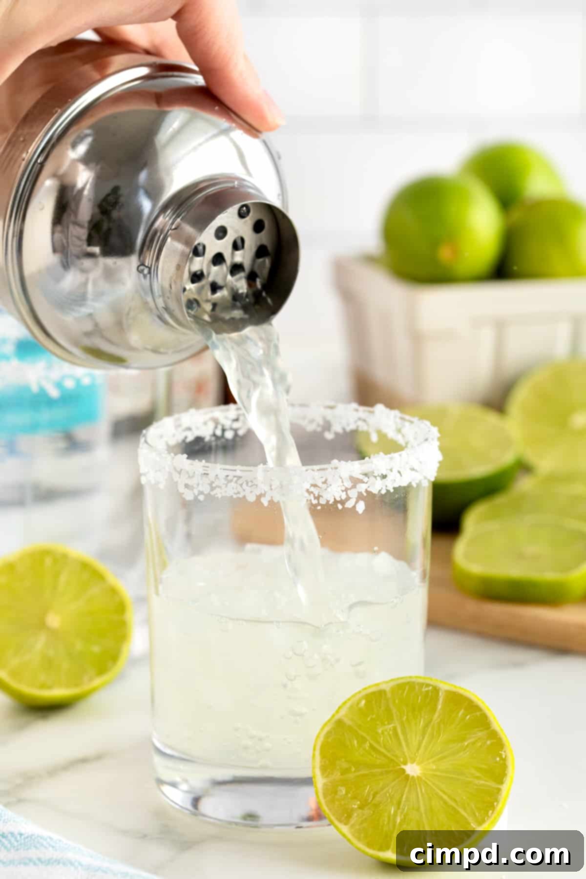 A salt rimmed glass being filled with margarita on a rimmed wooden serving tray. There is a lime wedge on the rim of the glass.