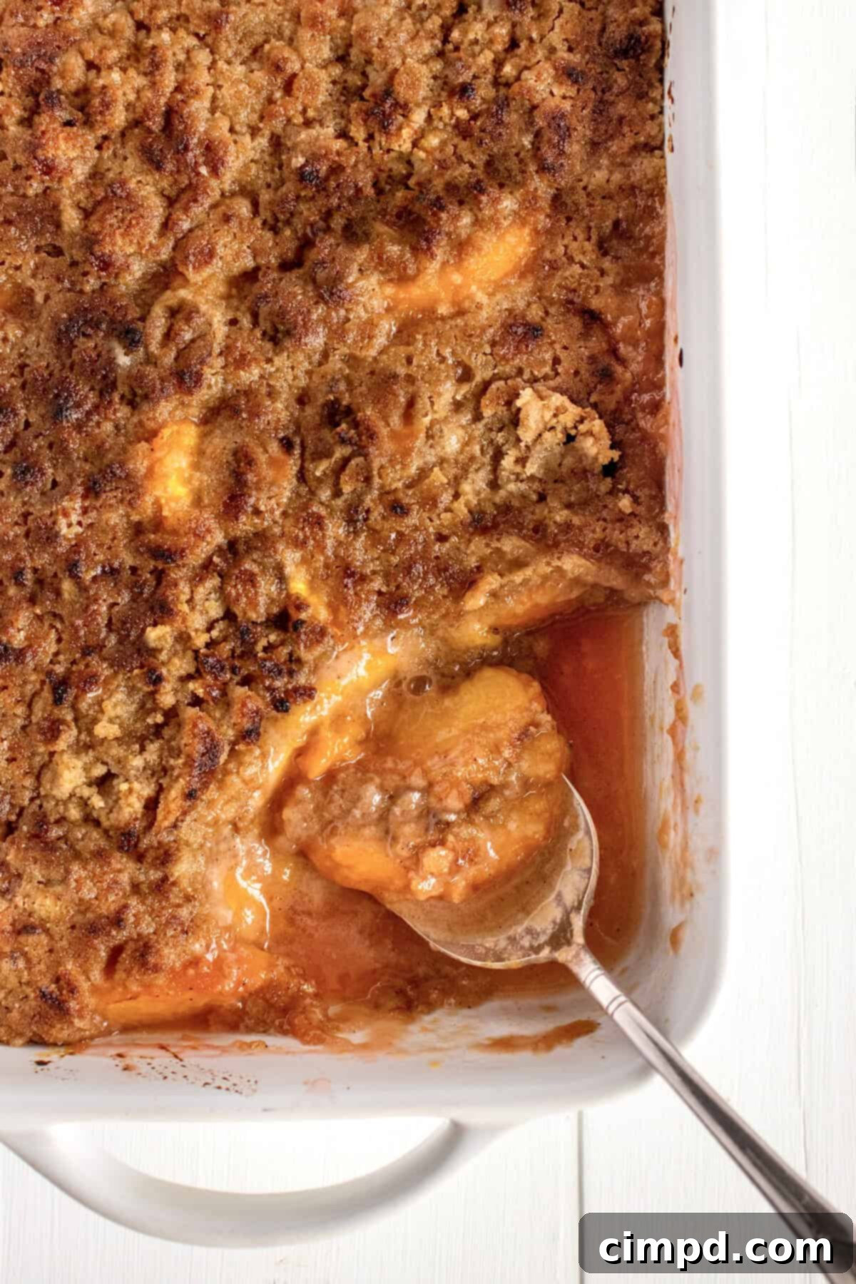 A baking dish of peach crumble with a serving removed and spoon laying in it.