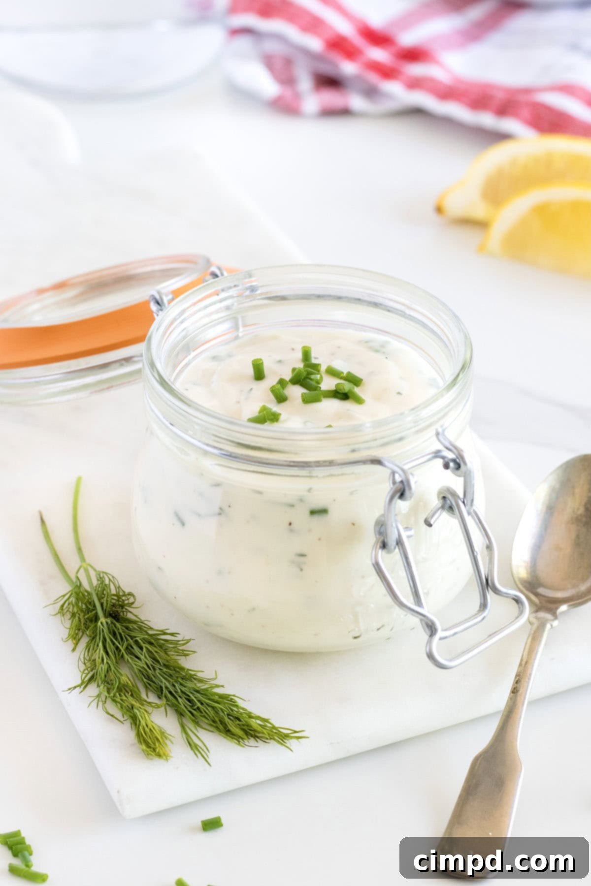 A glass jar of homemade ranch dressing on a white marble cutting board. There are fresh herbs to the bottom left.