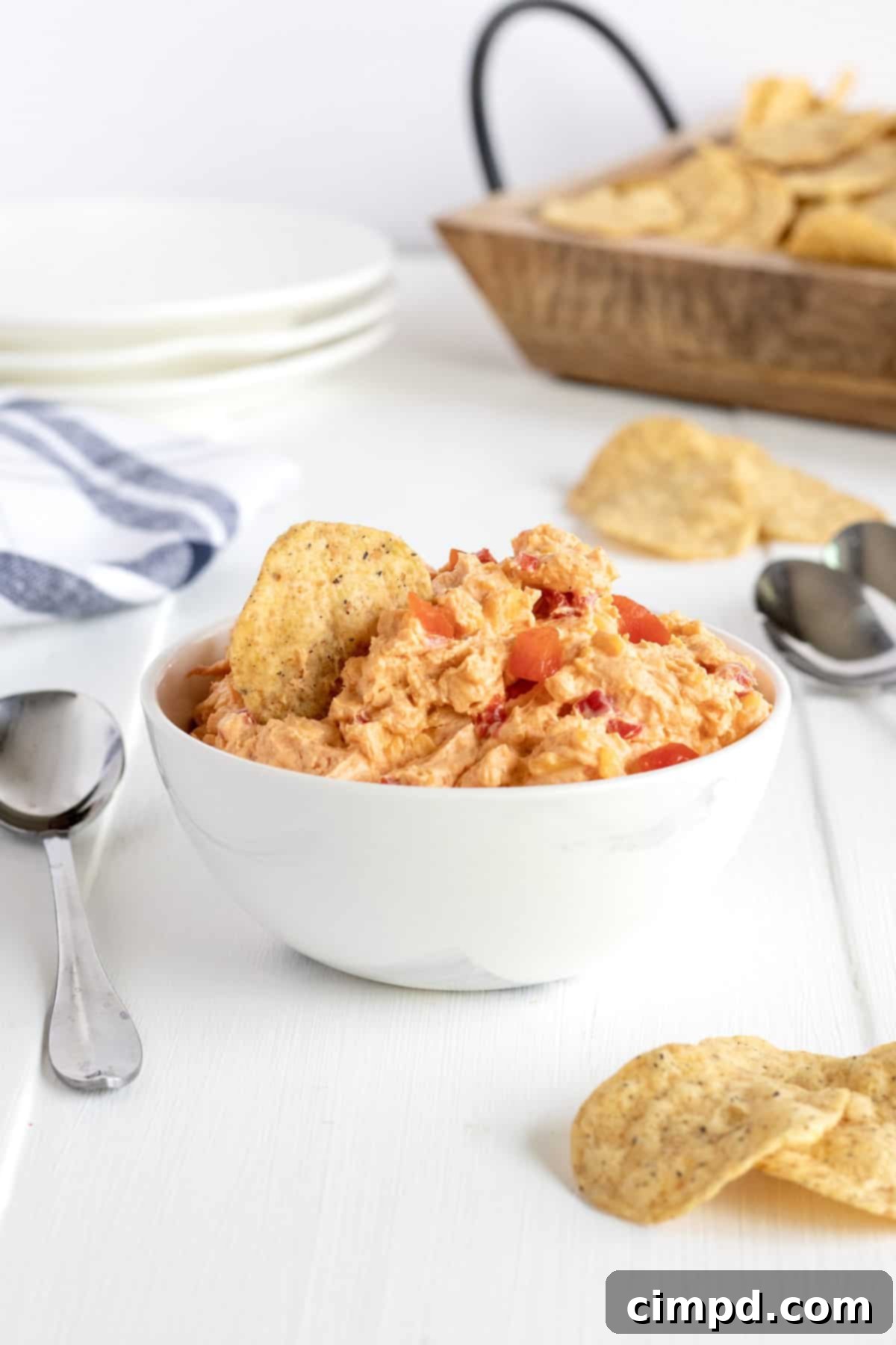 A pristine white serving bowl filled with creamy Pimento Cheese Dip, accompanied by a small spoon, resting on a clean white countertop, highlighting its smooth texture.