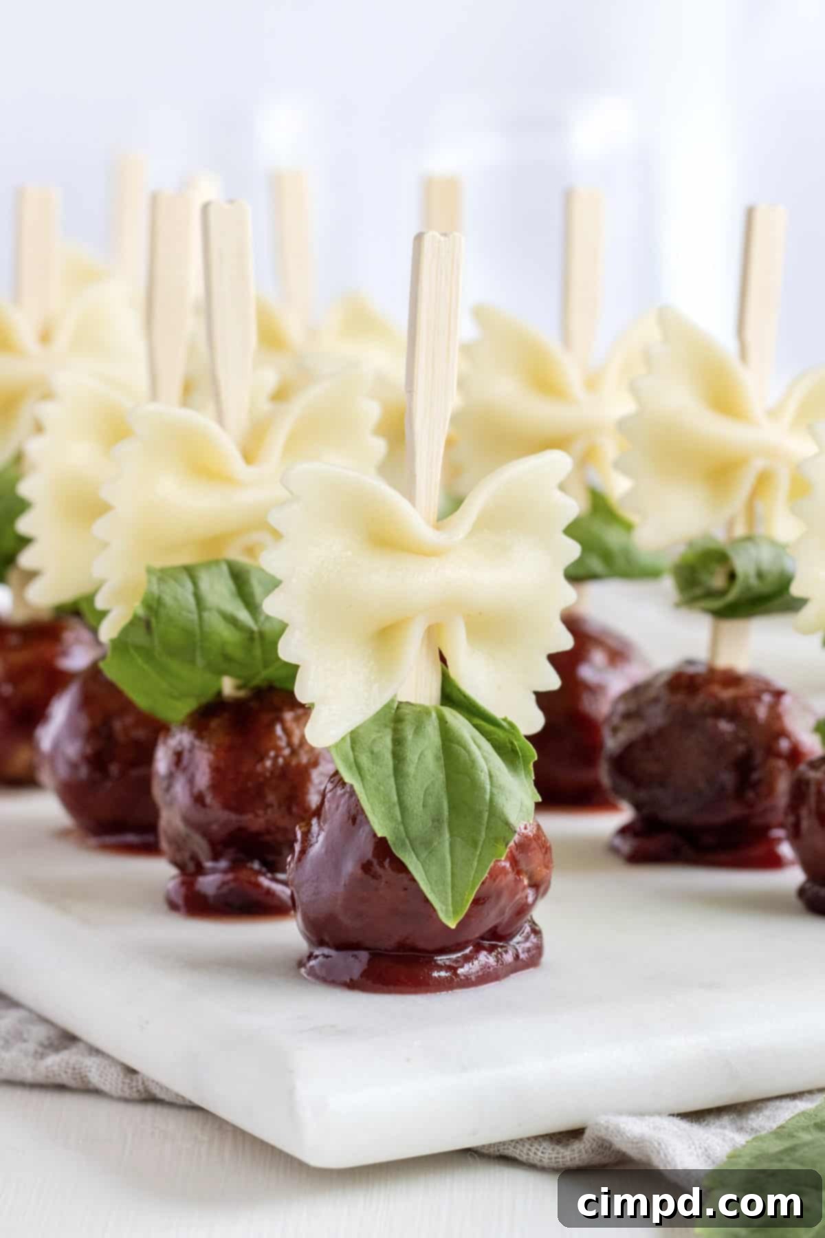 A beautifully arranged platter showcasing 12 wooden skewers, each adorned with perfectly cooked bowtie pasta and a glossy, saucy cocktail meatball, all resting on a pristine white marble serving tray. The presentation hints at an appetizer that's both elegant and utterly delicious.