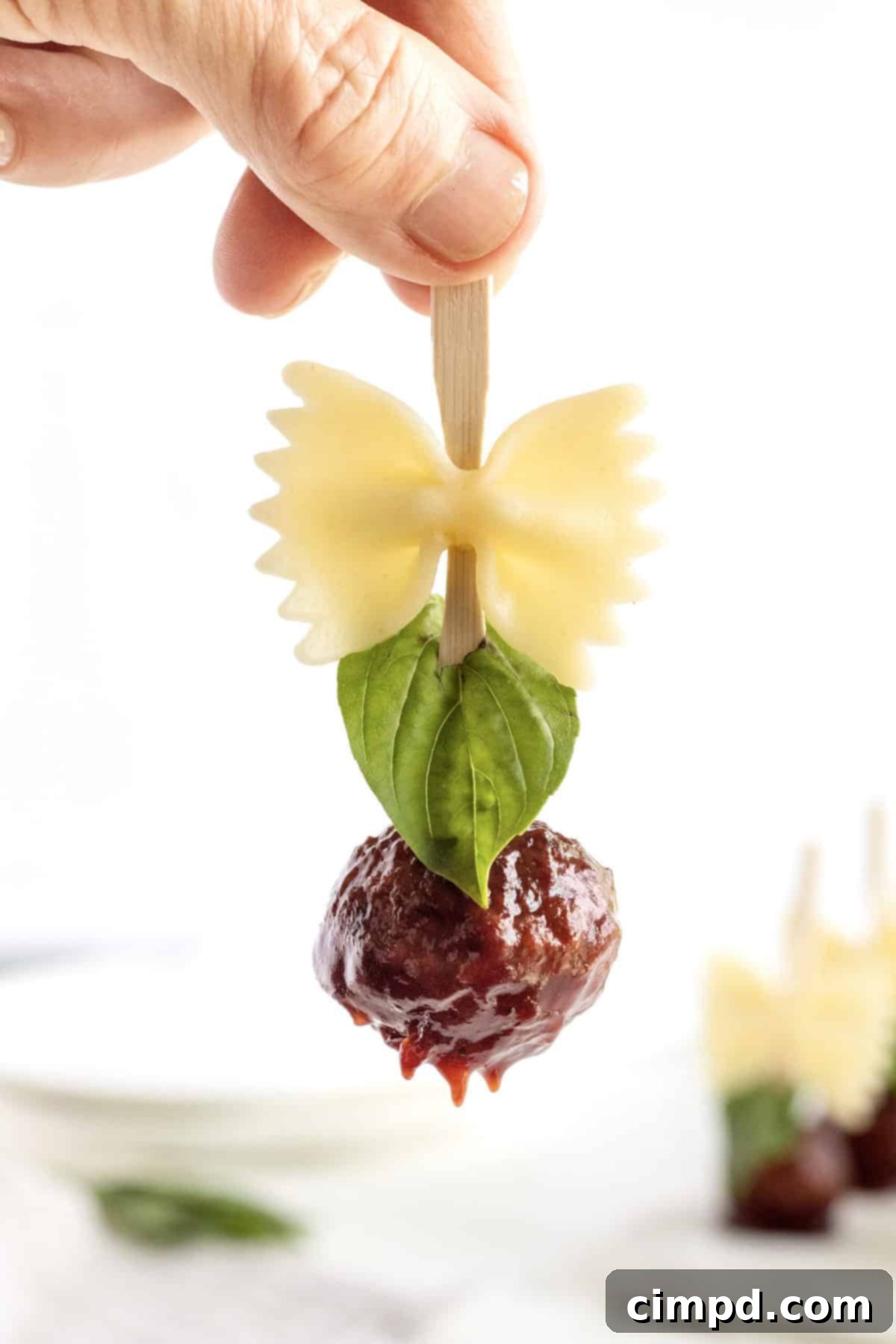 A close-up shot of a single wooden skewer, elegantly held between a thumb and forefinger. It features a piece of perfectly cooked bowtie pasta paired with a glistening, saucy meatball, highlighting the appetizing details of this charming appetizer.