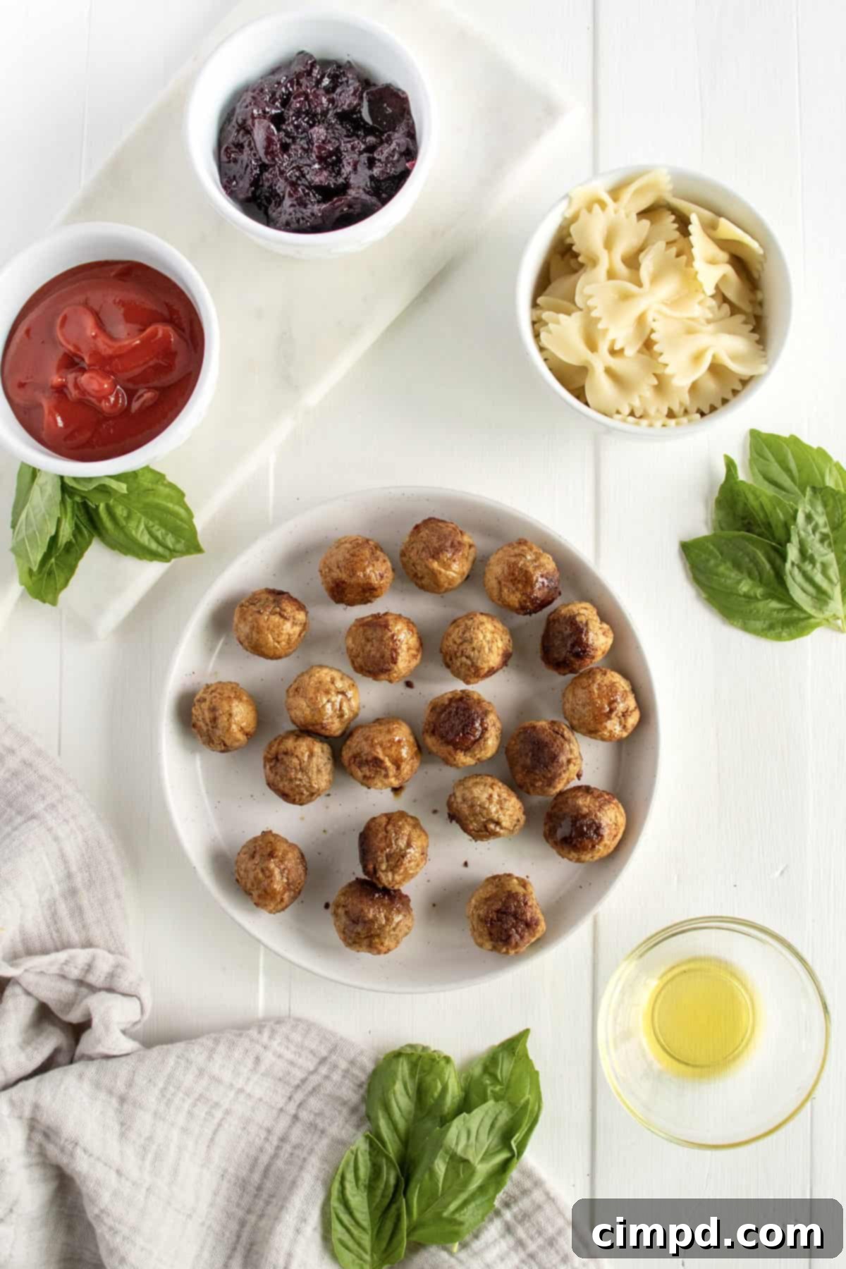 A flat lay photograph showcasing the key ingredients required for making Bowtie Pasta and Cocktail Meatball Skewers by The BakerMama. Neatly arranged are a bag of frozen meatballs, a bottle of ketchup, a jar of grape jelly, a box of bowtie pasta, a bottle of extra-virgin olive oil, and a bunch of fresh basil, all ready for preparation.
