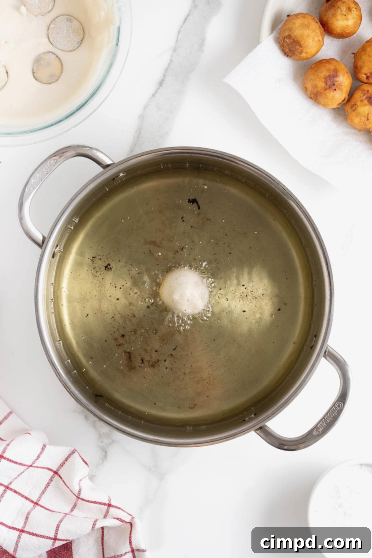 A single battered chocolate chip cookie dough ball is carefully lowered into a large frying pan filled with hot oil, beginning its transformation into a crispy, golden treat.