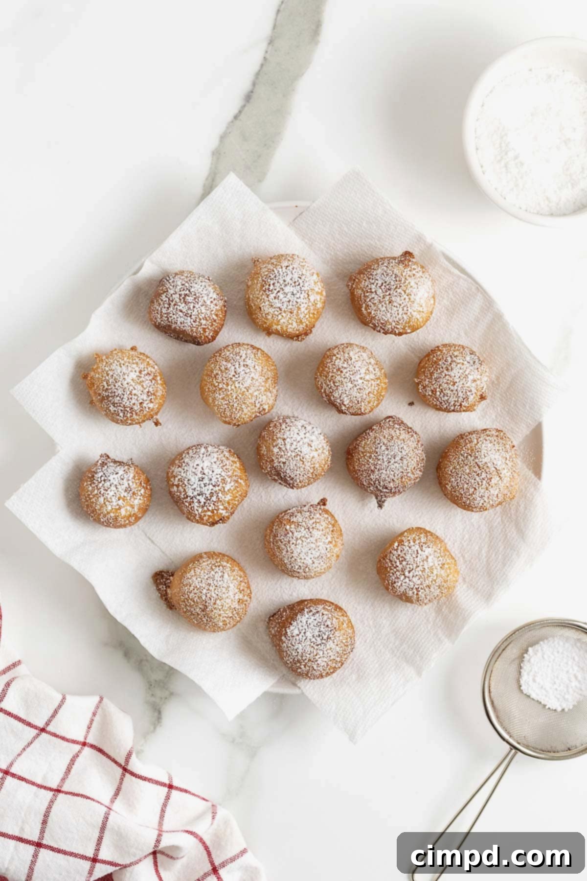 A close-up shot of freshly fried cookie dough bites, generously dusted with powdered sugar, resting on a white plate lined with paper towels, awaiting enjoyment.