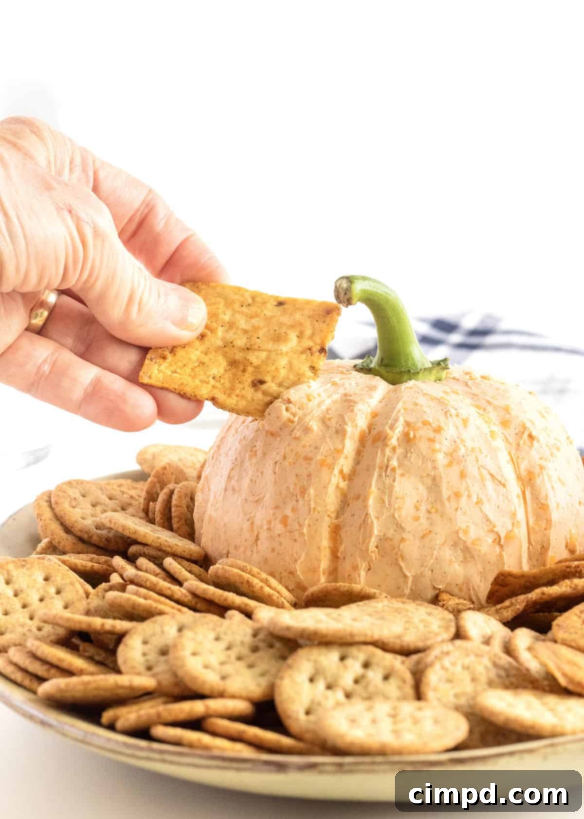 A cheese ball in the shape of a pumpkin surrounded by crackers on a large white serving plate. A hand is dipping a cracker into the cheeseball.