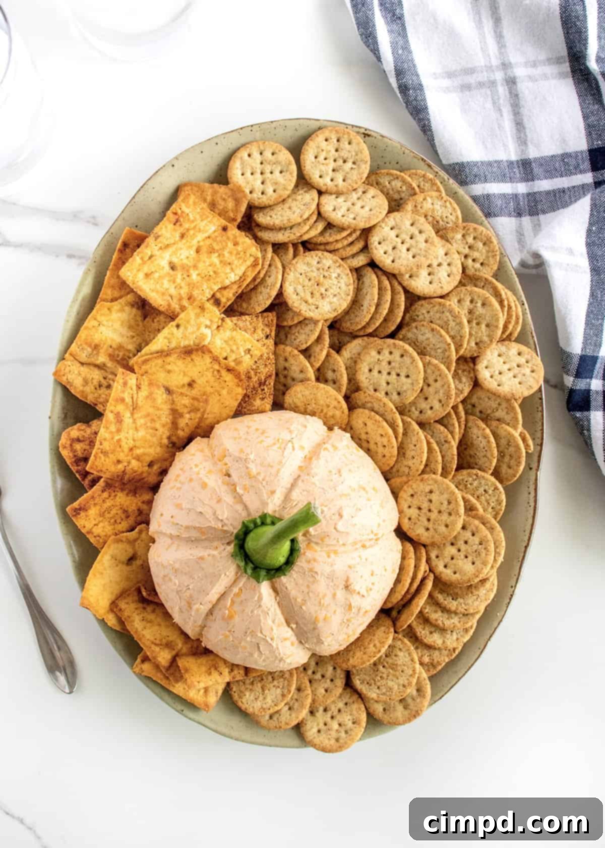 A cheese ball in the shape of a pumpkin surrounded by crackers on a large oval earthenware serving plate.