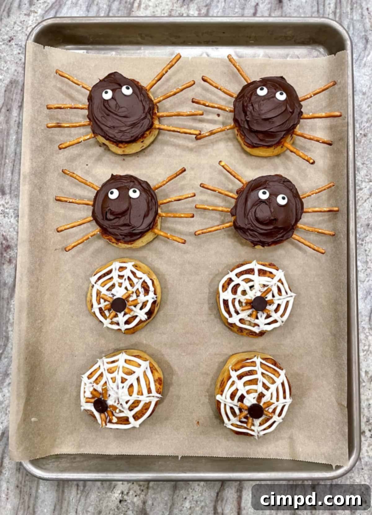Two cinnamon rolls decorated as a chocolate spider with pretzel legs and a white spiderweb with a chocolate chip spider.