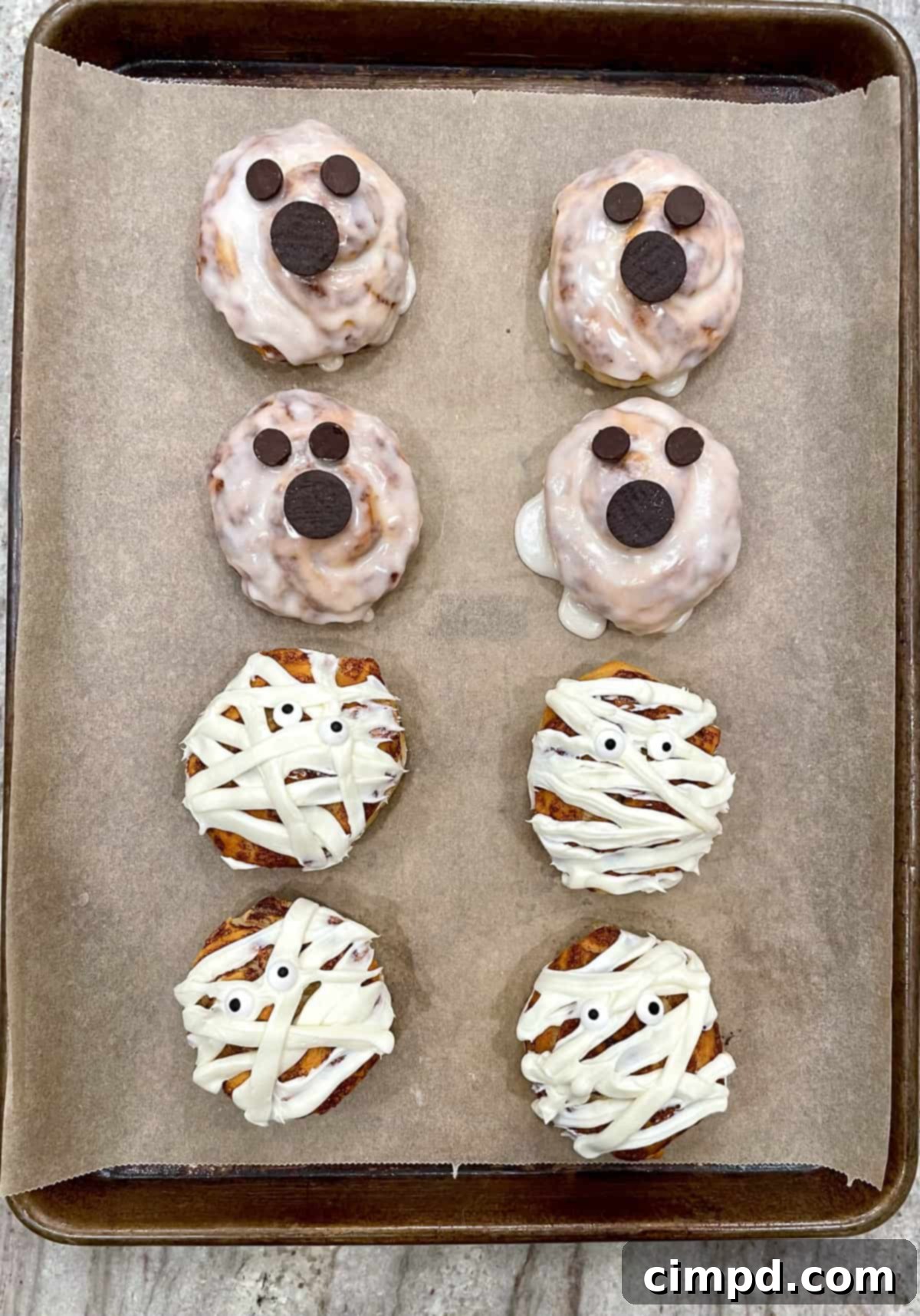 Two cinnamon rolls decorated as a white ghost with chocolate chip eyes and a white mummy with candy eyes.