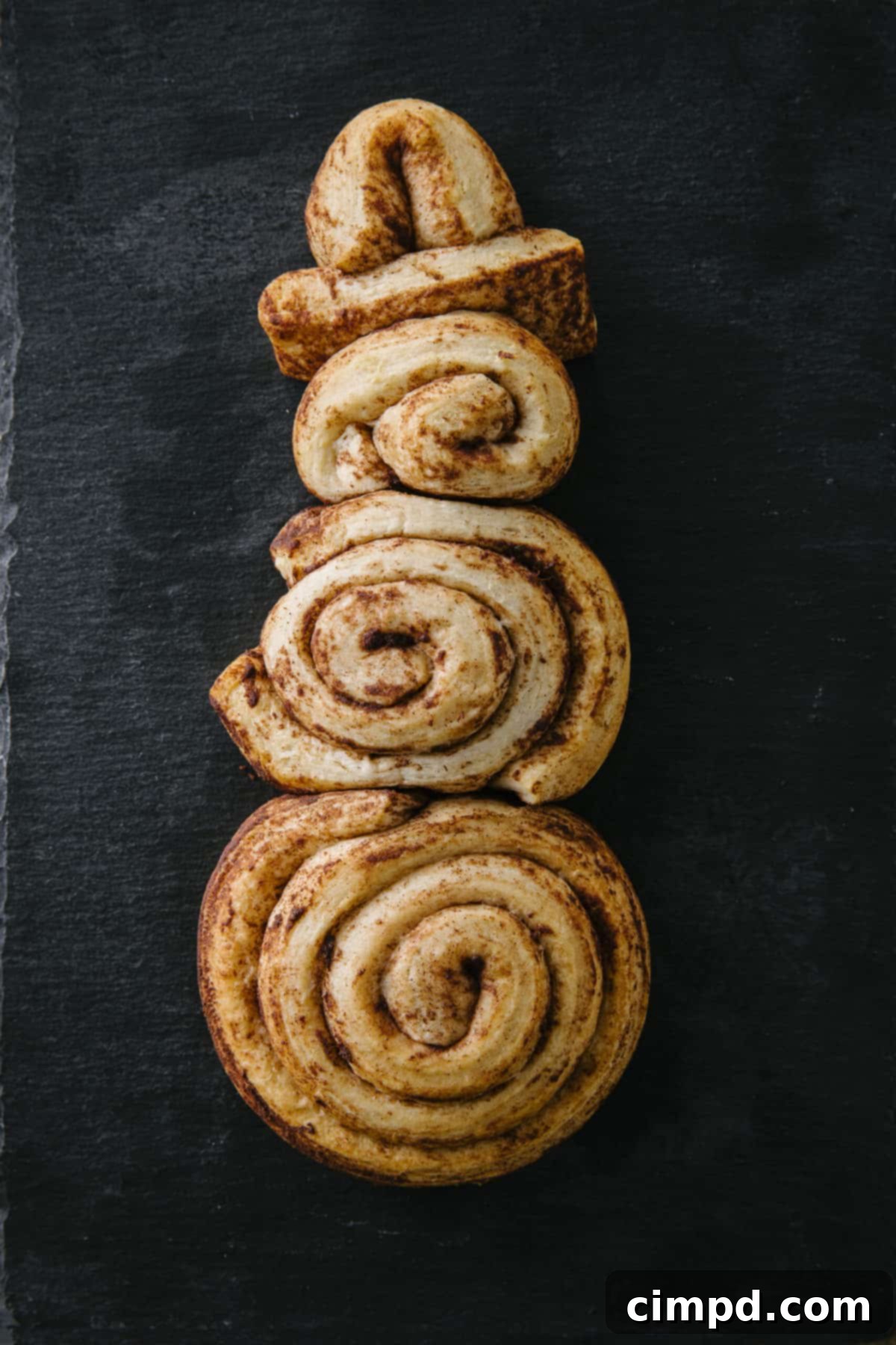 Frosty Cinnamon Creations 4 Cinnamon roll dough shaped into various snowman forms (snowman, snowkid, snowbaby) on a baking sheet, ready for the oven. Recipe by The BakerMama