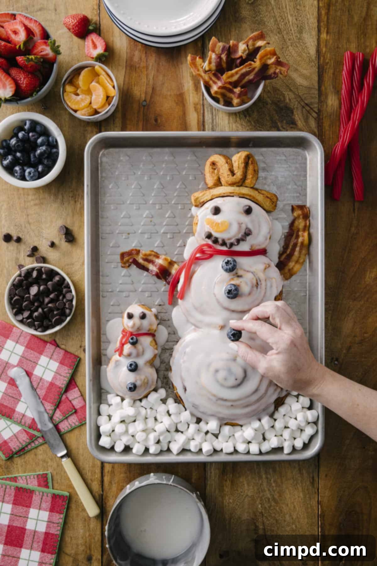 Frosty Cinnamon Creations 6 A festive spread of decorated Cinnamon Roll Snowmen (snowman, snowkid, snowbaby) with mini marshmallow 'snow,' blueberries, and bacon, ready for a holiday breakfast. Recipe by The BakerMama