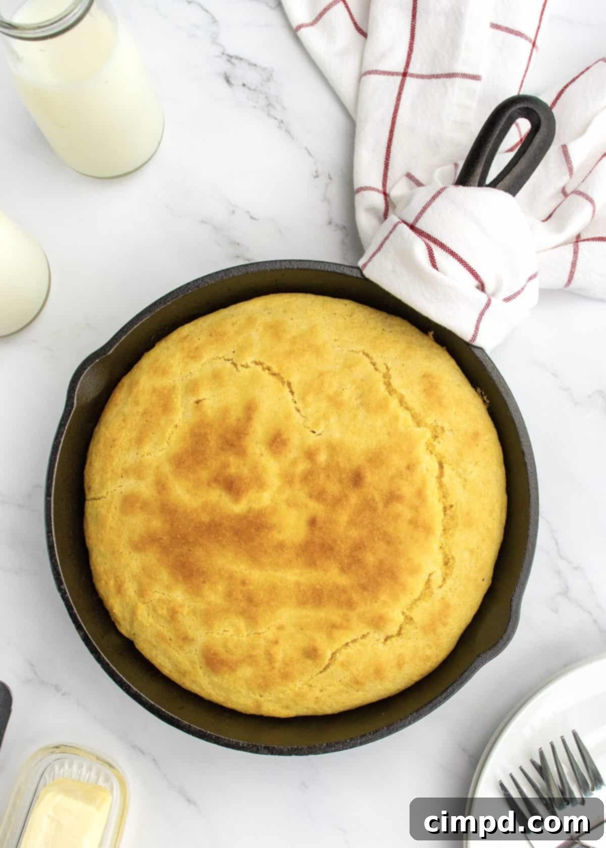 A cast iron skillet of cornbread on a white marble counter. There is a white towel with red stripes wrapped around the handle of the skillet.