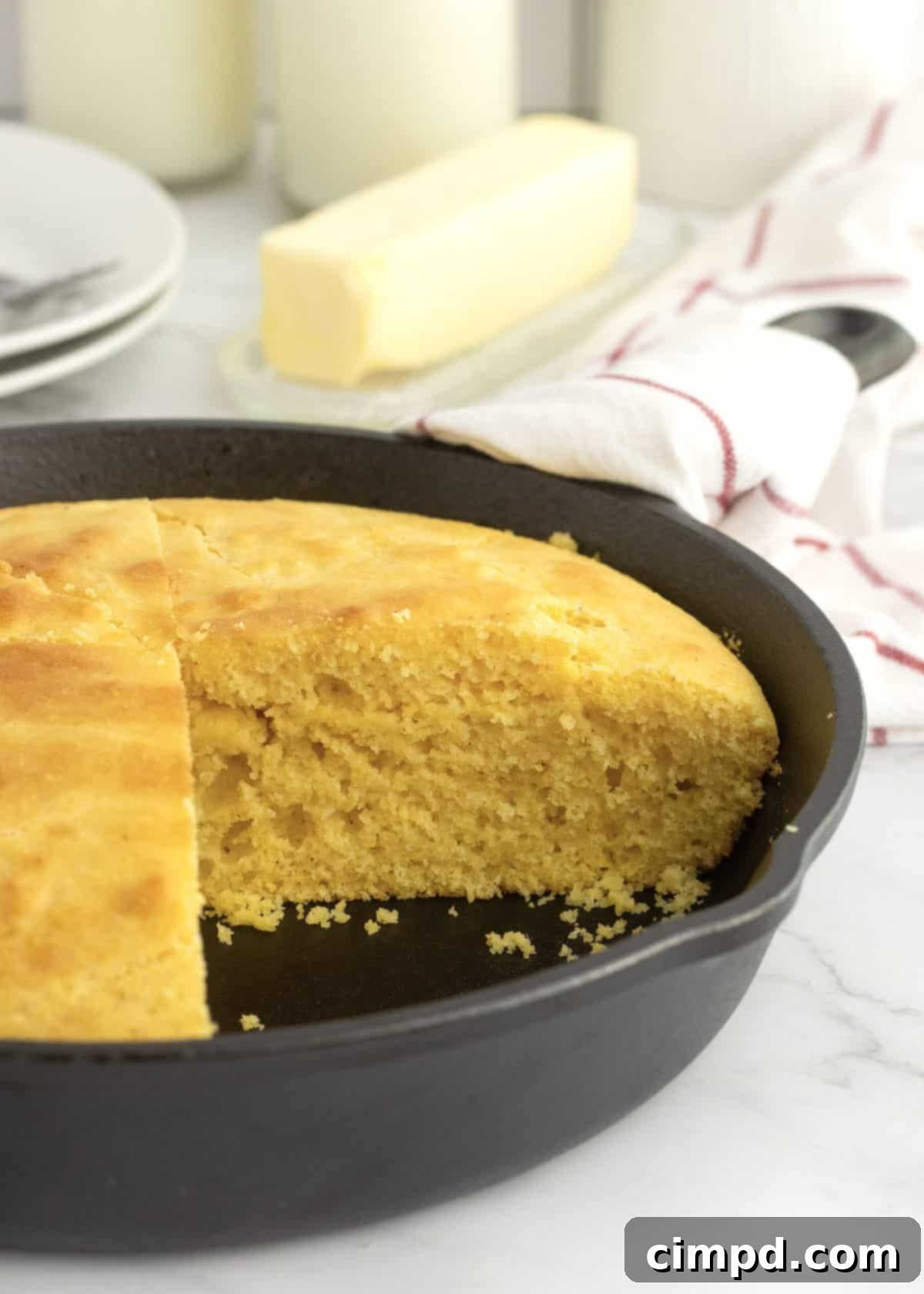 A cast iron skillet of cornbread on a white marble counter. There is a white towel with red stripes wrapped around the handle of the skillet. There is a piece missing from the cornbread.