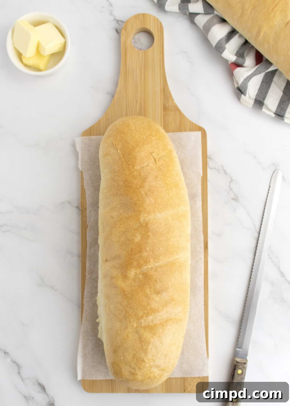 A freshly baked homemade French bread loaf resting on a rustic wooden cutting board, accompanied by a bread knife and a small dish of creamy butter.