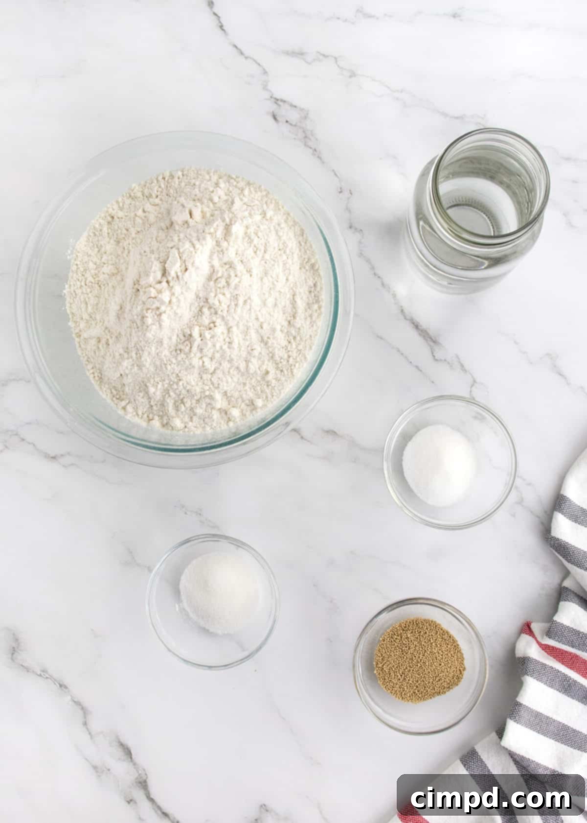 All five ingredients for homemade French bread – yeast, sugar, water, salt, and flour – are neatly arranged on a kitchen counter, ready for the baking process.